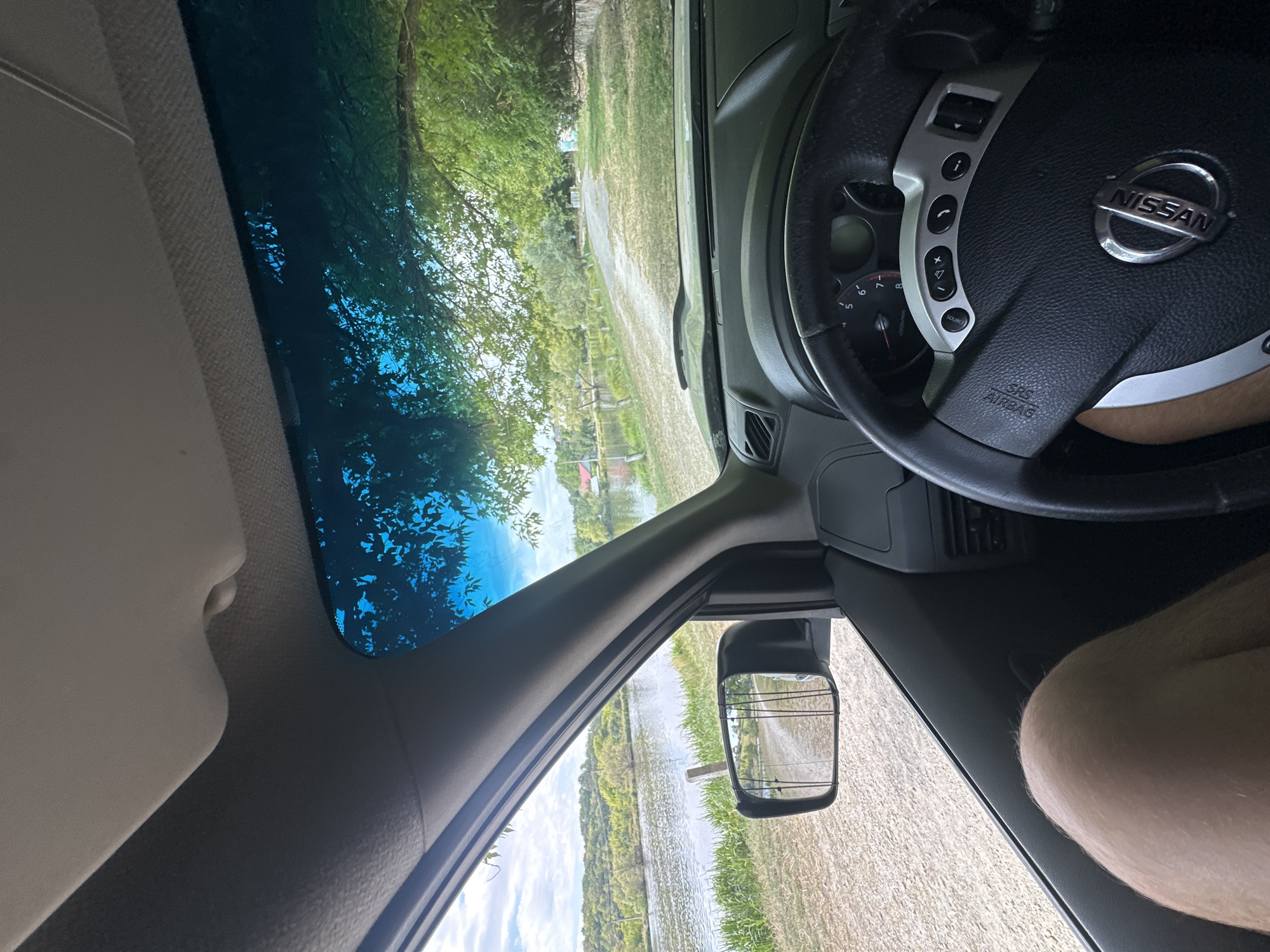 The image depicts the interior of a Nissan vehicle, with a view through the windshield and driver's side window. The driver's leg is visible in the lower left portion of the frame. The vehicle appears to be parked or moving slowly on a dirt road next to a body of water. The side mirror reflects a dirt road with several fishing rods. In the distance, there are trees, a few buildings with red roofs, and a body of water. The sky is partly cloudy. The overall scene suggests a rural or semi-rural setting, possibly a fishing spot. The image does not provide any specific information about the city of Makiv, Ukraine, or any events occurring there.