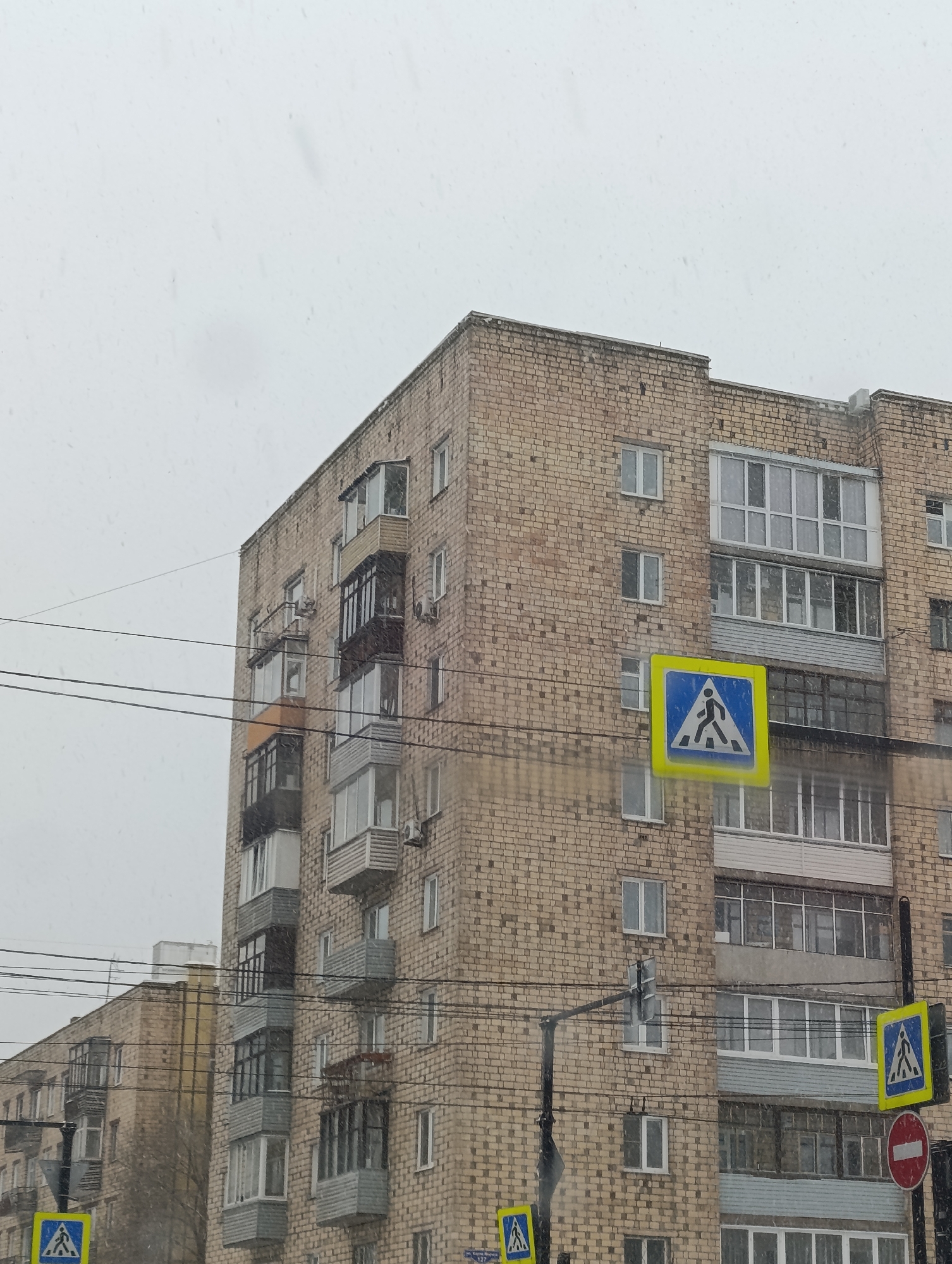 The photograph depicts a multi-story brick apartment building in Krasnoyarsk, Russia, taken on a cloudy, possibly snowy day. The building is constructed of light brown bricks and has numerous windows and balconies. Several pedestrian crossing signs are visible in the foreground, indicating a street intersection. Power lines run across the lower part of the image. The overall scene conveys a typical urban landscape in Russia during a cold weather season.