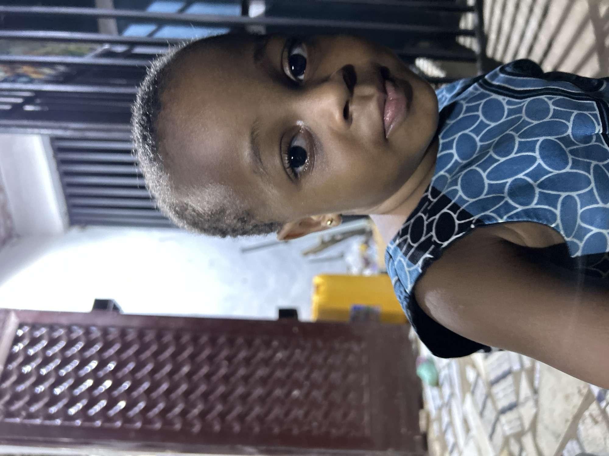 A close-up portrait of a child with dark skin, short hair, and dark eyes is captured indoors. The child is wearing a blue and black patterned shirt. The background includes a dark metal structure with horizontal lines, a light-colored wall, and part of a brown latticed surface. The location is Ihiagwa, Nigeria.