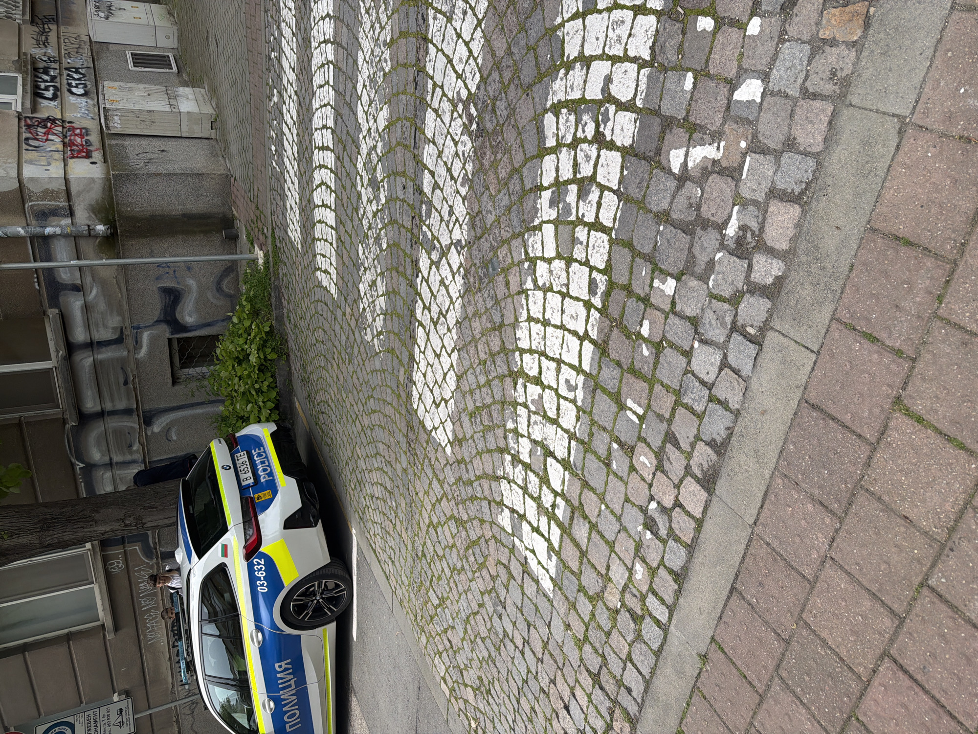 The image shows a Bulgarian police car parked next to a cobblestone pedestrian crosswalk in an urban setting. The car is white and blue, with "ПОЛИЦИЯ" (Police) written on its side, and a license plate number B 6536 TE. The car is parked on the edge of a gray asphalt road, next to a raised cobblestone section. The crosswalk consists of white stripes painted on the cobblestones. A sidewalk made of red brick pavers is visible in the foreground. Buildings with graffiti are in the background. The photo appears to have been taken in Varna, Bulgaria based on the license plate on the pole "ЖЕБЕН HAMEHT Места: 1 бр." meaning 'Reserved Places' and the phone number which begins with 052, the area code for Varna.