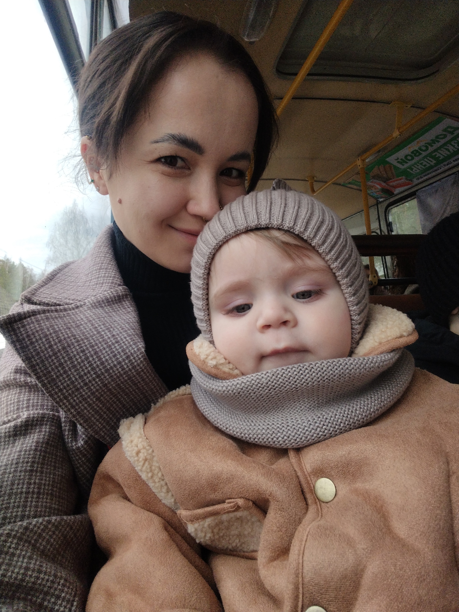 A woman and baby are in a bus or a vehicle that seems to be a form of public transportation. The woman is fair-skinned with short brown hair and a small mole on her left cheek. She wears a black turtleneck sweater and a brown and gray plaid coat. The baby, who is fair-skinned with light brown hair, is wearing a beige knitted hat and a light brown coat with a white wool collar and a gray knitted snood. The vehicle appears to be a bus, evident from the interior ceiling, metal bars, windows, and the advertising poster written in Russian visible above. The location might be somewhere in Noviy, Russia, given the signage, but confirmation requires a closer inspection. The scene is likely a bus interior; the individuals' interaction shows a mother taking care of her child.