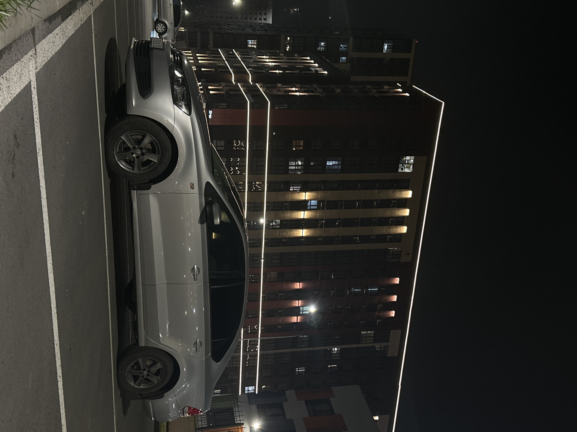 A silver Volkswagen Polo sedan is parked at night in a marked asphalt parking space within an urban residential complex in Rostov-on-Don, Russia. The vehicle is positioned in profile, facing left, with visible dark alloy wheels and tinted windows. In the background, a multi-story modern residential building, consistent with the architecture of the Red Aksay complex, features a dark facade accented with red-brown panels and prominent white LED strip lighting outlining its structure and horizontal bands. Vertical upward-facing lights illuminate portions of the building's facade. Numerous windows on the building are visible, with some emitting interior light. To the immediate right of the main building, another structure with orange and gray panels is partially visible behind a dark metal fence. A streetlight illuminates the area between the car and the buildings. Another vehicle is partially visible parked further back on the left. The sky is dark, indicating nighttime.