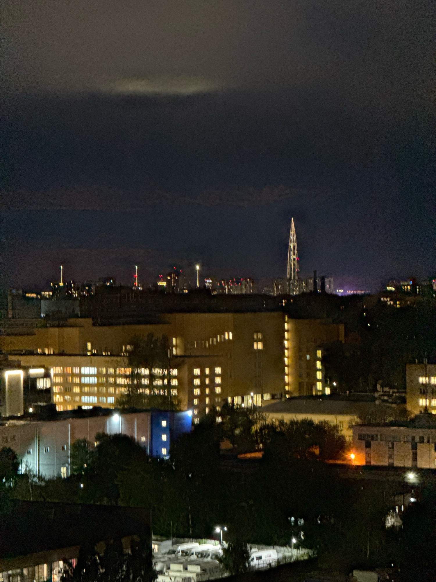 The image is a nighttime aerial view of a city skyline in Saint Petersburg, Russia, dominated by the illuminated Lakhta Center skyscraper, a tall, needle-like structure dominating the distant horizon. The foreground and midground are filled with various buildings, many of which have illuminated windows, suggesting residential or office spaces. Trees and darker, less distinct structures occupy the lower portions of the image. The sky is dark with some faint clouds, and there are several other distant city lights visible, including what appear to be radio towers or tall structures with lights. The overall impression is one of urban sprawl at night, with a prominent architectural landmark.