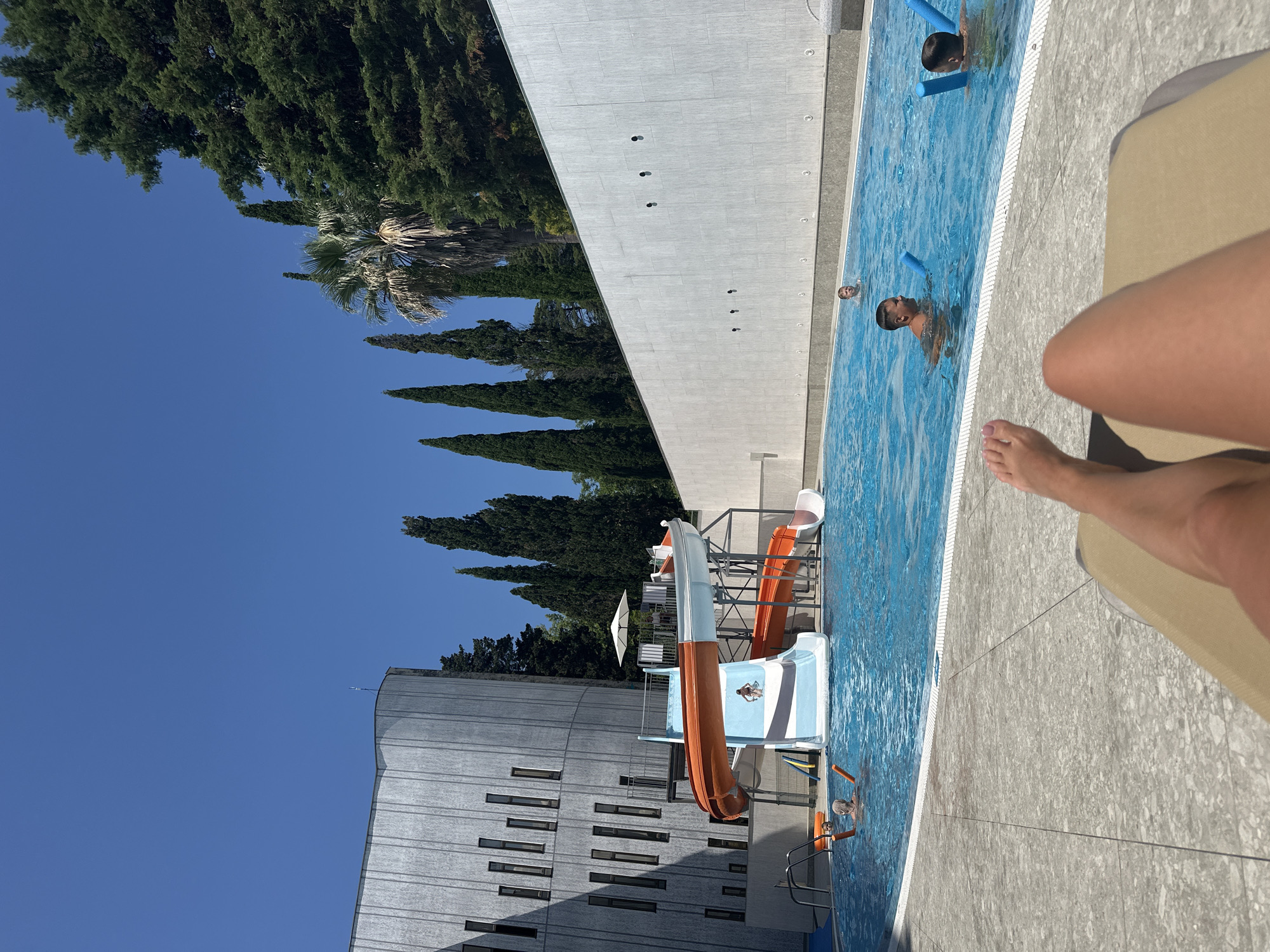 The image depicts an outdoor swimming pool area at a resort in Sochi, Russia, on a clear, sunny day. The pool is filled with bright blue water and features a water slide complex with orange and white slides. Several people are visible in the pool: two boys are playing with blue foam noodles, one man is in the water, and a child is in the distance. In the foreground, the legs and feet of a person relaxing on a lounge chair are visible, along with the chair's headrest.
Behind the pool, a modern, cylindrical building with a textured gray facade and numerous vertical windows is present, as is a long, white, textured wall. A variety of tall, slender trees, including cypresses and a palm tree, line the background, suggesting a lush, subtropical environment characteristic of Sochi. The overall scene conveys a relaxed atmosphere of a vacation resort.