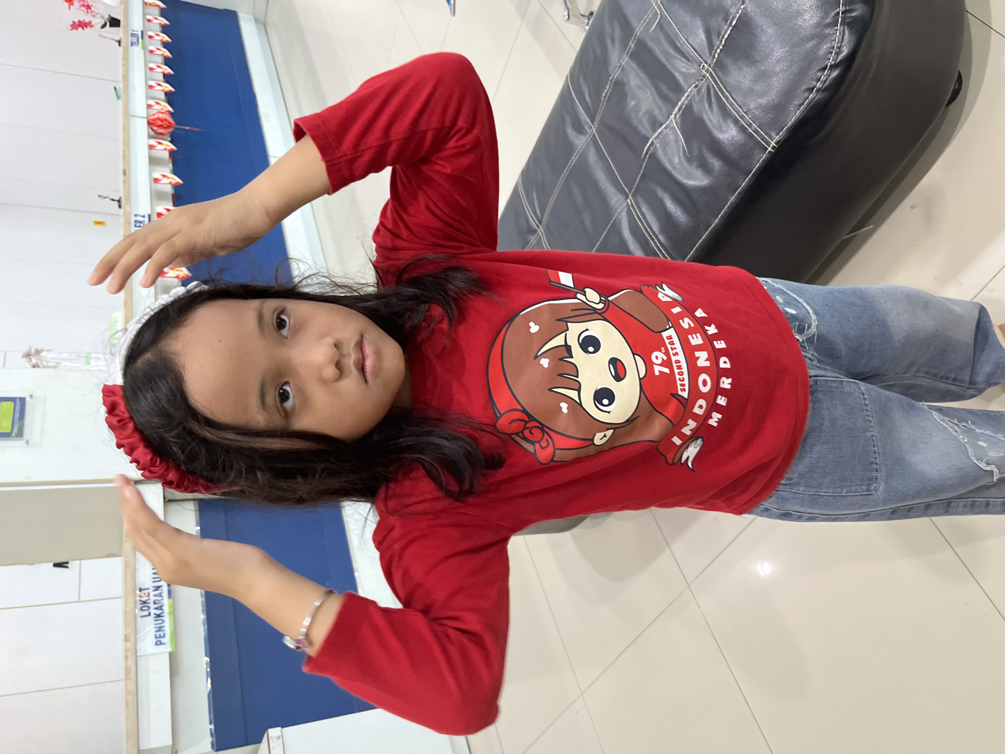 A young girl wearing a red long-sleeved shirt with a cartoon graphic and denim jeans stands in what appears to be an indoor waiting area. The graphic on her shirt depicts a cartoon character holding an Indonesian flag and text that includes "INDONESIA MERDEKA." She is wearing a red and white headband and has her hands on the headband, slightly touching her head. In the background, there is a counter with signs and decorations, suggesting a public service area. To the right, a black leather bench is partially visible. The flooring is tiled. The overall scene suggests a celebratory or patriotic theme, possibly related to Indonesian Independence Day.