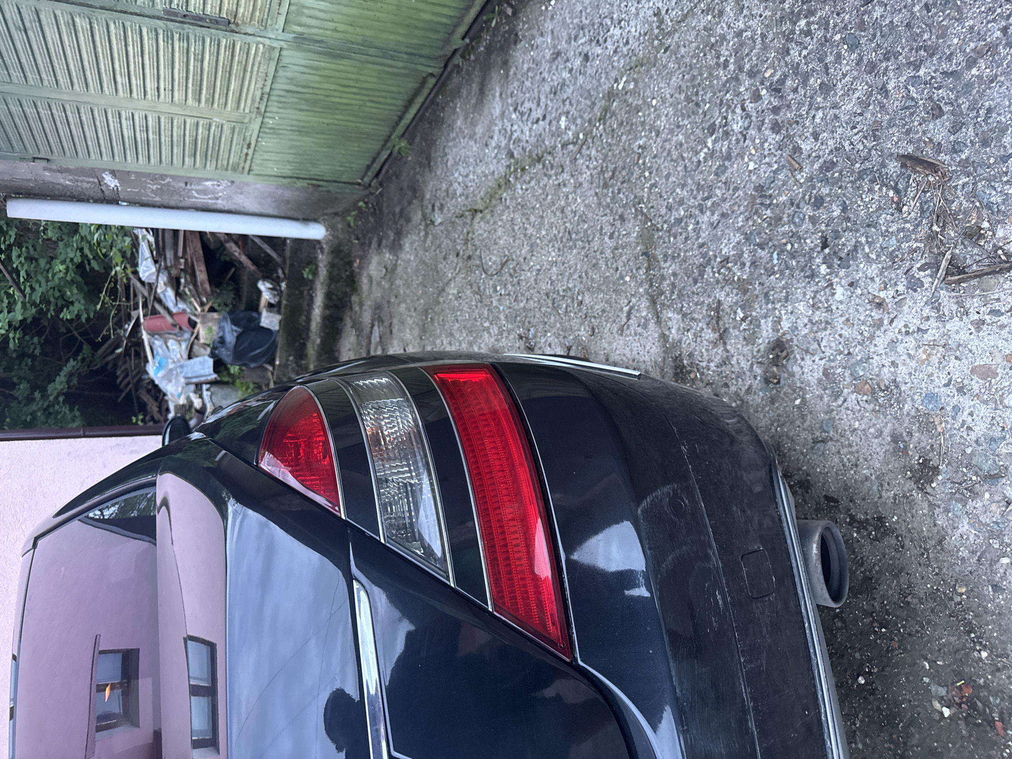 The image shows the rear of a dark-colored car parked on a concrete surface. The car has a distinctive taillight design with red and white sections, along with a visible exhaust pipe. In the background, there is a green corrugated metal garage door and a pile of discarded items. A pink building is partially visible on the left side of the frame. The location is likely Vratsa, Bulgaria, though specific landmarks or features to confirm this definitively are absent.