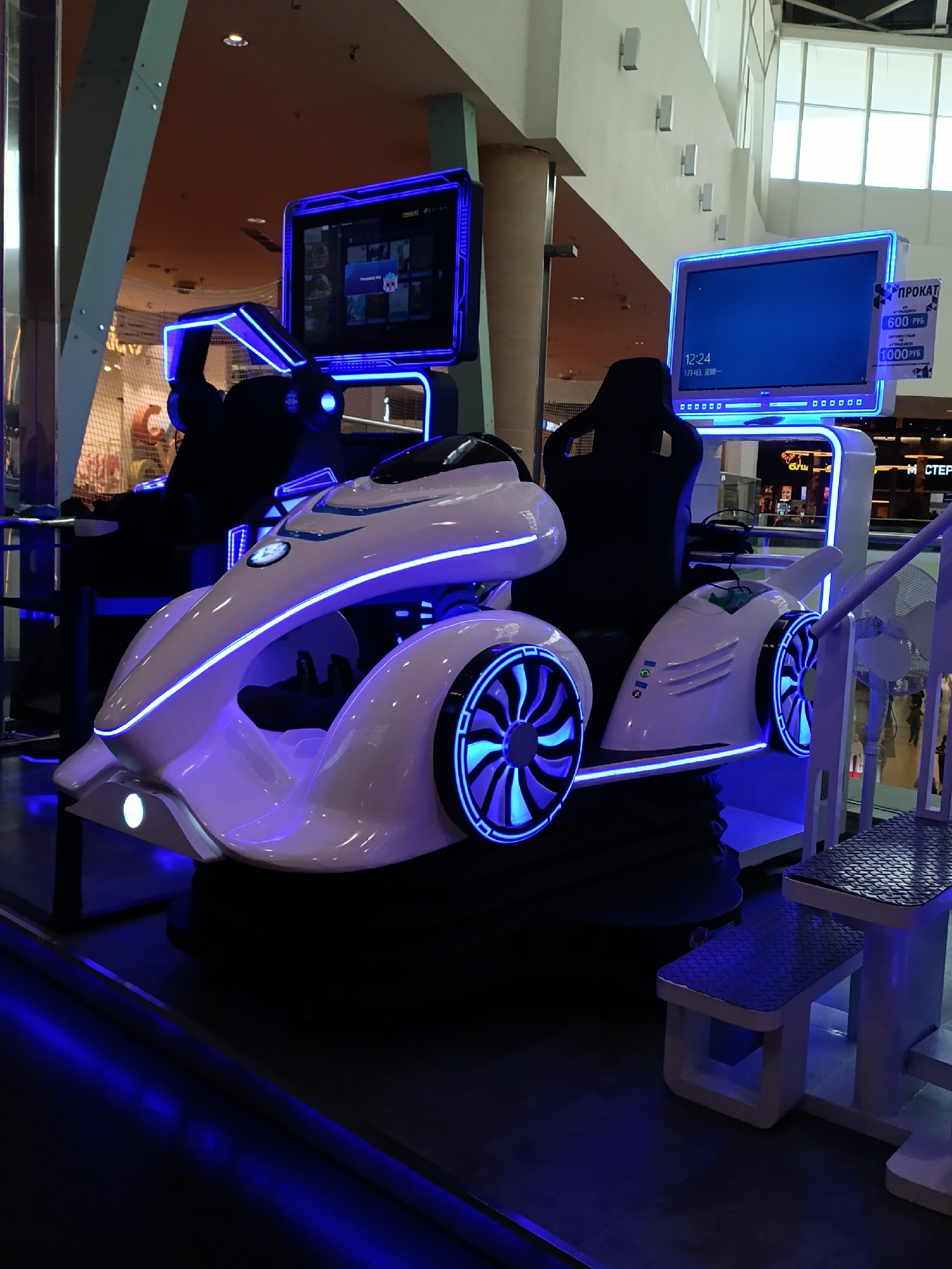 The image shows two virtual reality ride simulators in what appears to be a shopping mall in Krasnoyarsk, Russia. The simulators resemble futuristic race cars, featuring white exteriors with blue neon accents and black seats. Each simulator has a large display screen above the seat and another to the side, one displaying a menu and the other showing time and price information. The platforms for the simulators have metal steps, and the area is lit with blue ambient light, creating a futuristic, arcade-like atmosphere.