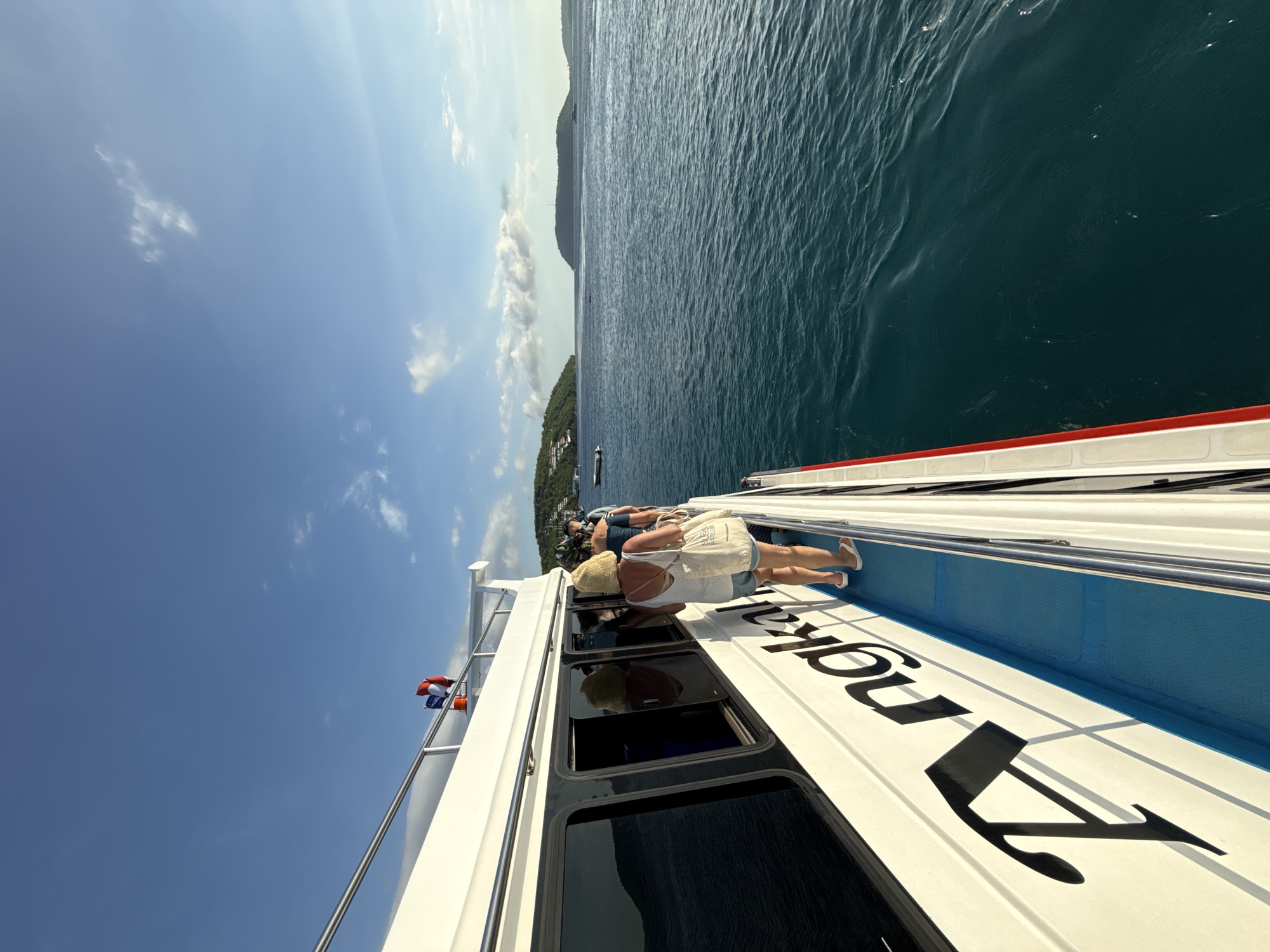 From the deck of a white boat labeled "A Naka" in black lettering, a group of people are observing the surrounding landscape. One woman with a straw hat and a white tank top is leaning over the side of the boat to look out the window. The boat is on a body of water with an island visible in the distance. The sky is clear with some clouds. Based on the presence of "Kampung Toyapakeh, Indonesia" in the prompt, it can be inferred that this scene is taking place in this location.