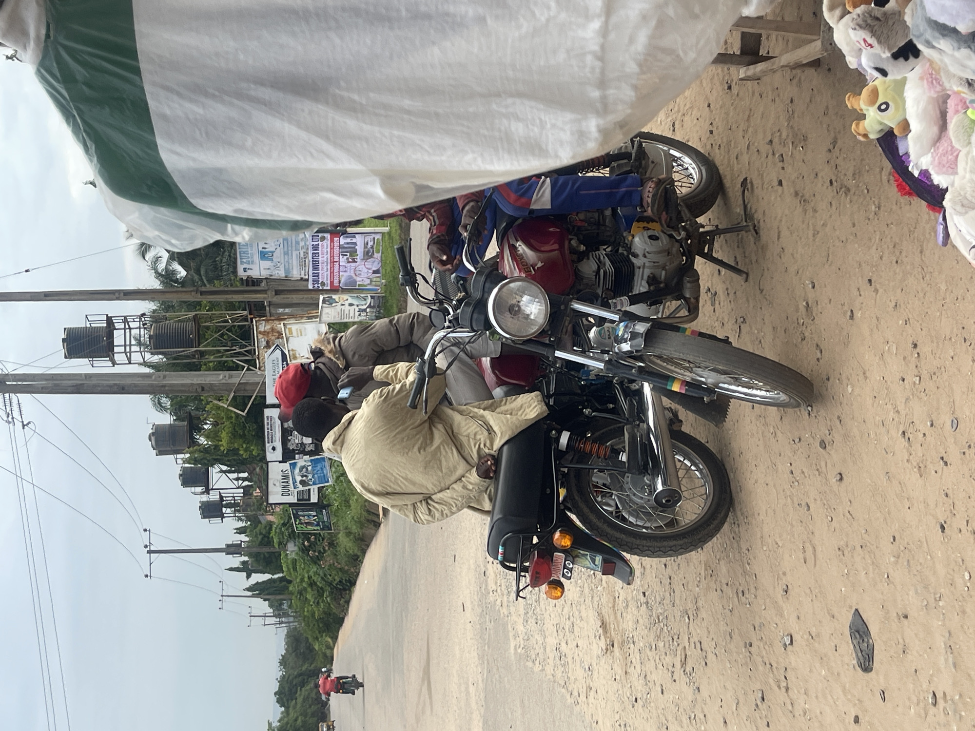 In Abuja, Nigeria, a dusty roadside scene unfolds with several individuals and motorcycles. Two men, one wearing a light-colored jacket and the other a red cap, are seated on a red motorcycle. The man in the jacket has his hands clasped behind his back, while the man with the red cap is talking on a mobile phone. Another individual, wearing a hooded jacket and blue pants with red and white stripes, is also on the motorcycle, positioned behind the other two. A third motorcycle, ridden by a person in a red shirt, is seen in the distance on the road. The environment features a paved road, a dirt shoulder with scattered stones, and overhead power lines supported by utility poles. In the background, several large water tanks are visible on raised structures, alongside trees and advertising billboards. A white and green tarp or sheeting is draped over a wooden frame in the foreground, partially obscuring a collection of stuffed animals and other items laid out for sale on the ground. The sky is overcast, suggesting cloudy weather.