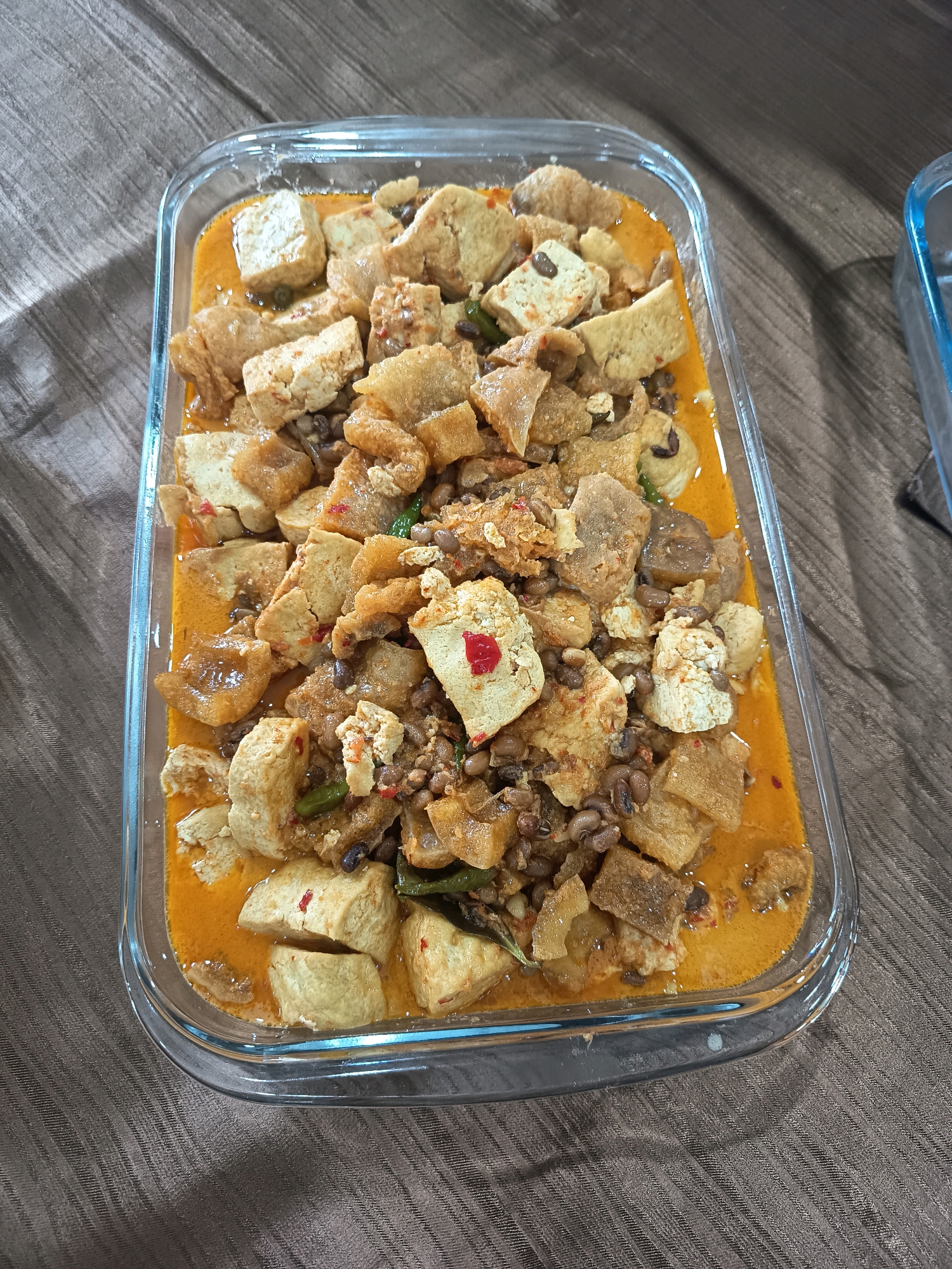A glass rectangular dish filled with a hearty Indonesian dish is placed on a textured brown tablecloth. The dish contains cubed tofu, pieces of fried pork skin, and black-eyed peas in a rich, orange curry sauce. Small pieces of chili and green chilies are visible within the dish. The lighting suggests an indoor setting, possibly a home kitchen or dining area, in Bogor, Indonesia. The dish is centered in the frame, with the tablecloth extending outwards. Another glass dish is partially visible to the right of the frame.