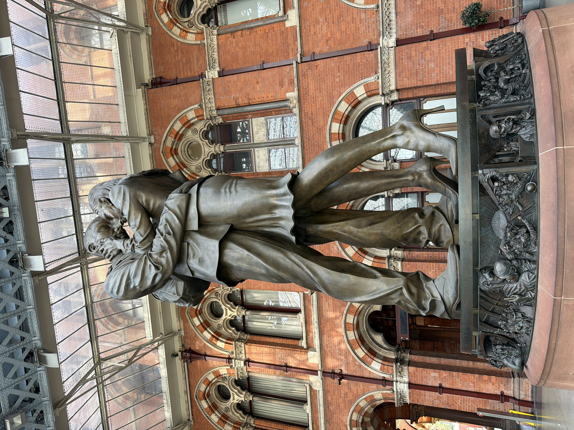 The image shows a statue titled "The Meeting Place" located in St. Pancras International train station in London, UK. The statue depicts a couple embracing, likely symbolizing reunions and farewells that occur at the station. The figures are cast in bronze and stand on a plinth adorned with carved panels illustrating various scenes. The backdrop includes the station's Victorian architecture, with red brick walls, ornate windows, and a glass roof structure.