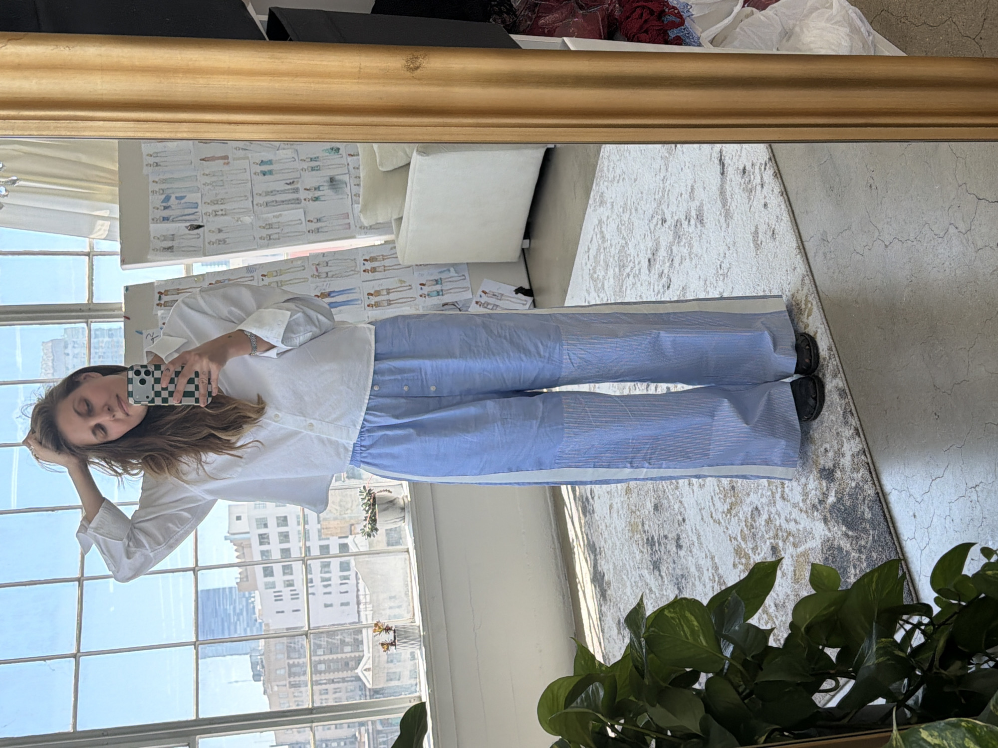 A medium-shot shows a woman in a large framed mirror, posing in a white button-up shirt and light-blue striped pants with a white side-stripe. She holds her phone, clad in a green and white checkerboard case, up to take a selfie. Behind her is a large, multi-pane window. Outside the window, buildings are visible, indicating that this scene is likely taking place in a high-rise.
In the background, several details are evident. A pile of red, white, and black clothing is visible behind the mirror's top edge. In the mirror’s reflection, there are sketches or design drawings taped to a wall, as well as a white couch. A shaggy grey and white rug partially covers the floor. There is a plant, with dark-green glossy leaves, in the lower right corner of the scene.
Based on the architecture seen through the window, this location is likely in a high-rise building within the city of Los Angeles, California, United States of America.