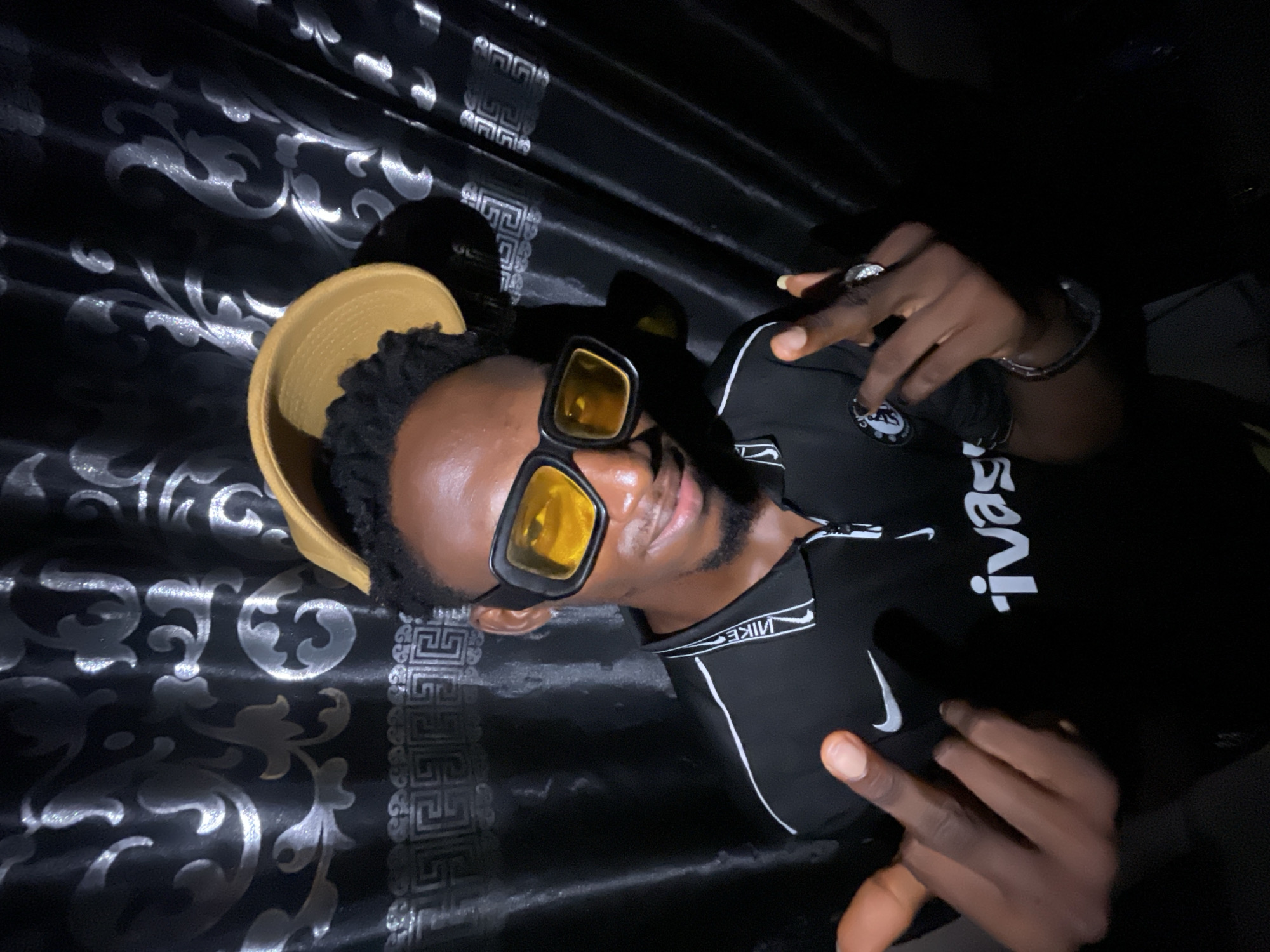 A person wearing a black Nike shirt, yellow-tinted sunglasses, and a yellow bucket hat is making a peace sign. The background appears to be black fabric with a silver pattern. The location in Lokoja, Nigeria is not specified in the image and there is no information to infer where it was shot.