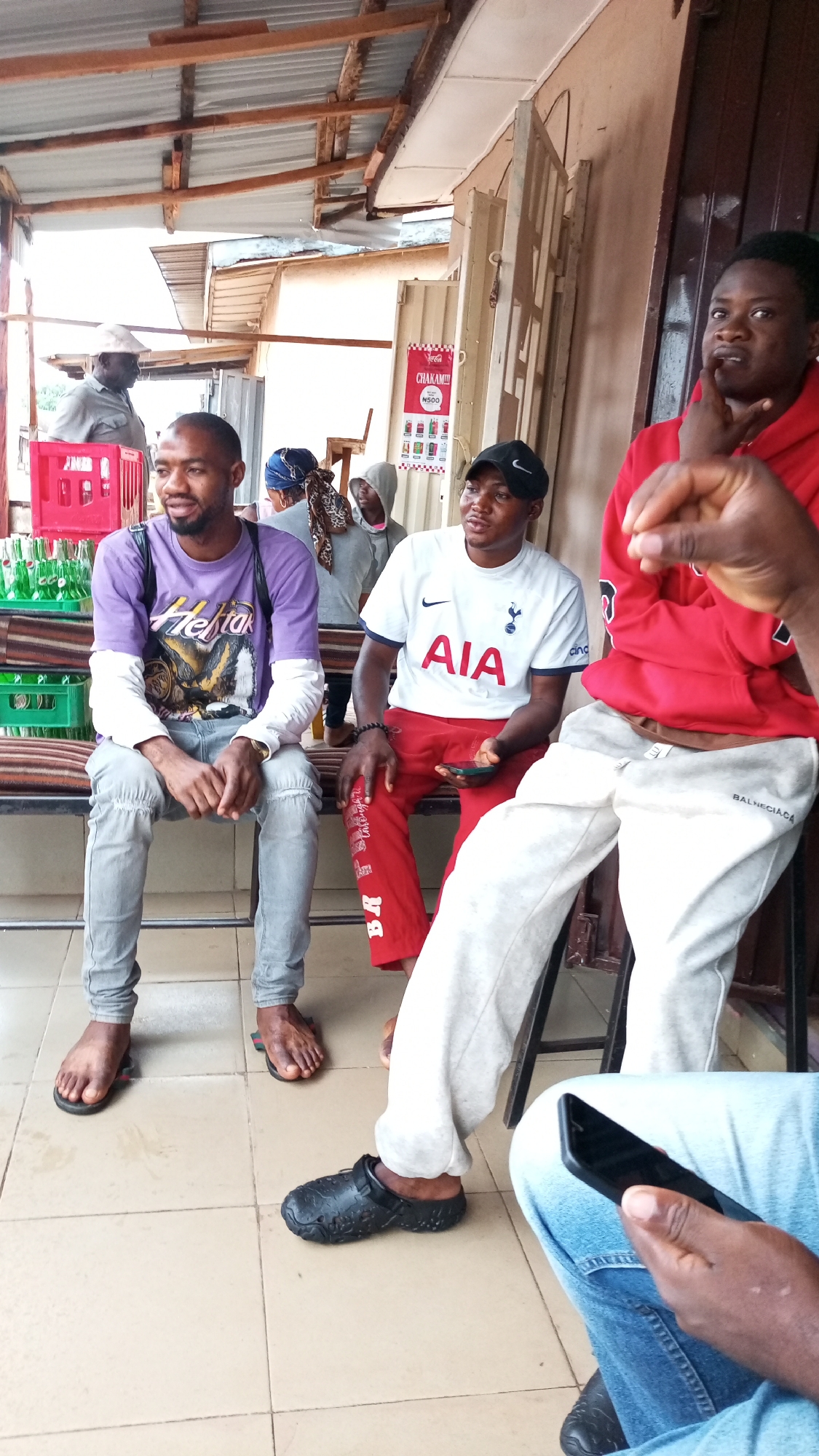 **Scene:** A casual outdoor gathering in Sabon Tasha, Nigeria.
**People:**
* Three men are prominently featured in the foreground. One is wearing a purple graphic tee, jeans, and sandals. Another wears a Tottenham Hotspur jersey, red pants, and sandals. The third man, partially obscured, wears a red hoodie and light gray Balenciaga sweatpants.
* Other individuals are visible in the background, including an older man in a hat and several figures partially concealed within the building.
**Objects:**
* Plastic crates of soft drinks (likely Coca-Cola) are stacked near the seated men.
* The man in the red hoodie is partially obscured by a stool.
* A hand holding a smartphone is visible in the lower right corner.
* A sign advertising drinks (possibly Coca-Cola) with prices is on the building.
* The floor is tiled with light brown squares.
**Actions/Interactions:**
* The men are seated and appear to be engaged in conversation or observing their surroundings.
* One man has his hand raised in a gesture.
* One of the men is holding a mobile phone.
**Environment:**
* The setting is an open-air space, potentially a small shop or roadside establishment.
* A corrugated metal roof covers the area.
* The building appears to be simple in construction, with some visible signs of wear.
* There is a white metal window frame.
