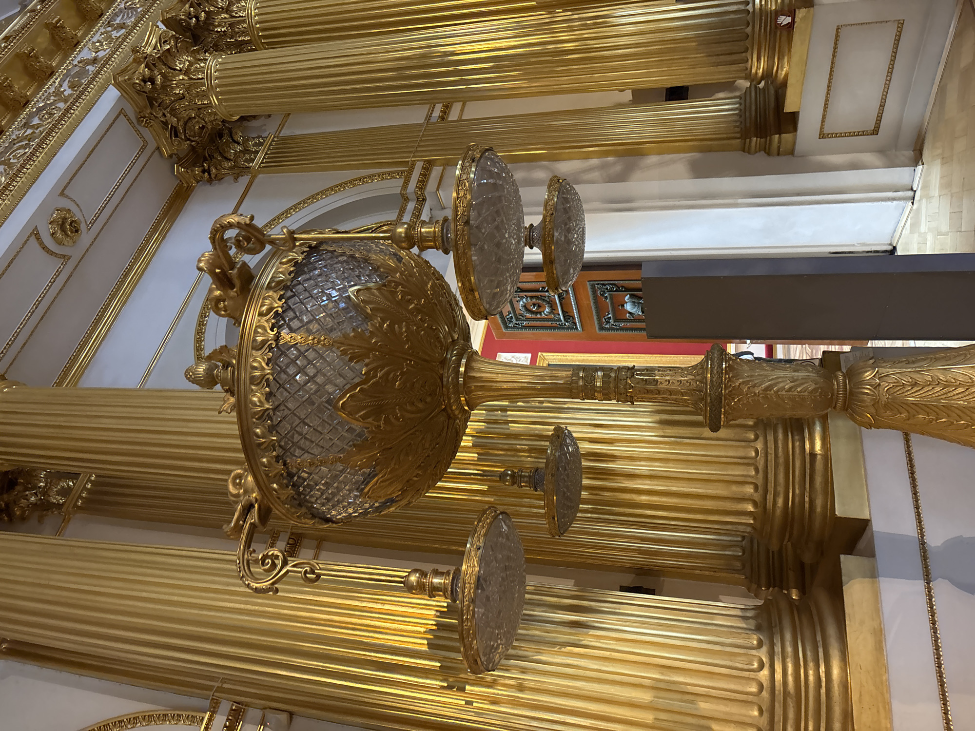 An ornate, gilded chandelier with a crystal bowl is displayed in a grand interior space with tall, fluted golden columns. Several smaller, glass sconces hang from the main fixture. The floor is made of light-colored parquet. The background features additional golden columns and architectural details, as well as a glimpse of a doorway with red walls and framed artwork. A dark gray rectangular object stands in the foreground. The lighting is bright and reflects off the metallic surfaces, creating a dazzling effect.