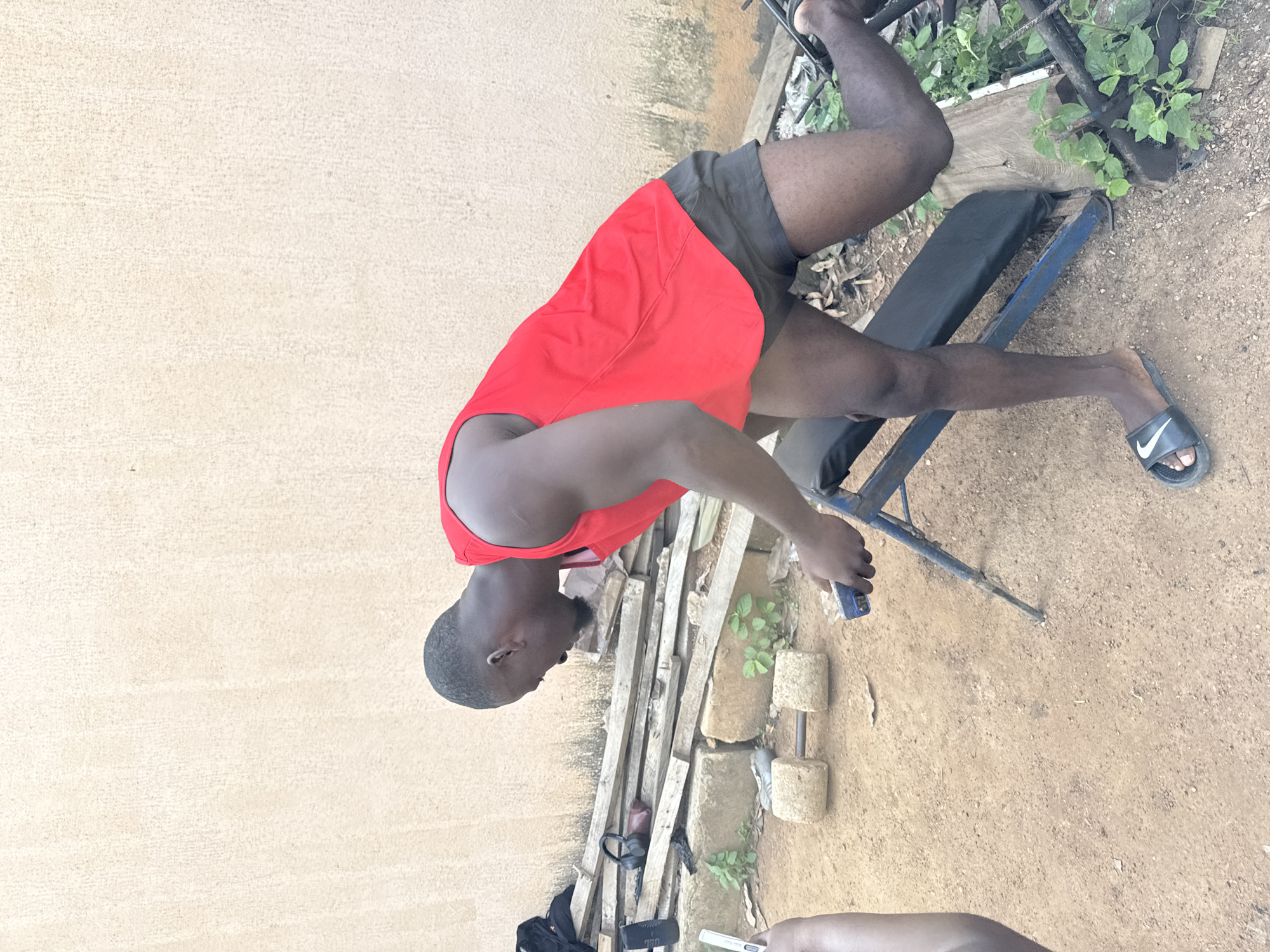 In Akwanga, Nigeria, a dark-skinned man wearing a red tank top and dark shorts is performing a workout outdoors. He is leaning over a blue weight bench, with his right leg extended backward and his left leg bent. His right hand is holding a blue object, possibly a phone. In the background, a wall made of light brown plaster forms the backdrop, with discarded wooden planks and large stones at its base. A handmade dumbbell with two large stone weights and a metal bar lies on the sandy ground to the left of the man. Green vegetation is visible around the workout area.