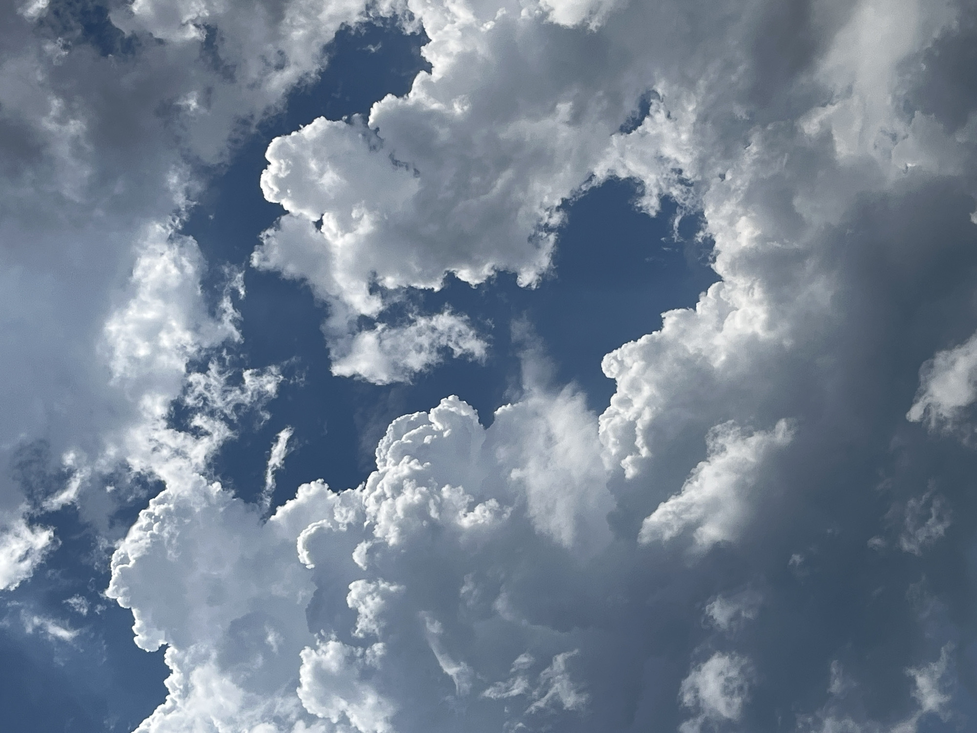 The image is a close-up, upward-looking shot of a cloudy blue sky. Large, fluffy white cumulus clouds dominate the frame, with pockets of deep blue sky visible between them. Some of the clouds have darker, more shadowed undersides, suggesting depth and texture. The lighting indicates a bright day, likely daytime, with sunlight illuminating the tops of the clouds. There are no people, structures, or land visible in the image; it is solely focused on the atmospheric conditions. The location is specified as Golden Beach, Greece, but this context is not visually evident from the image itself.