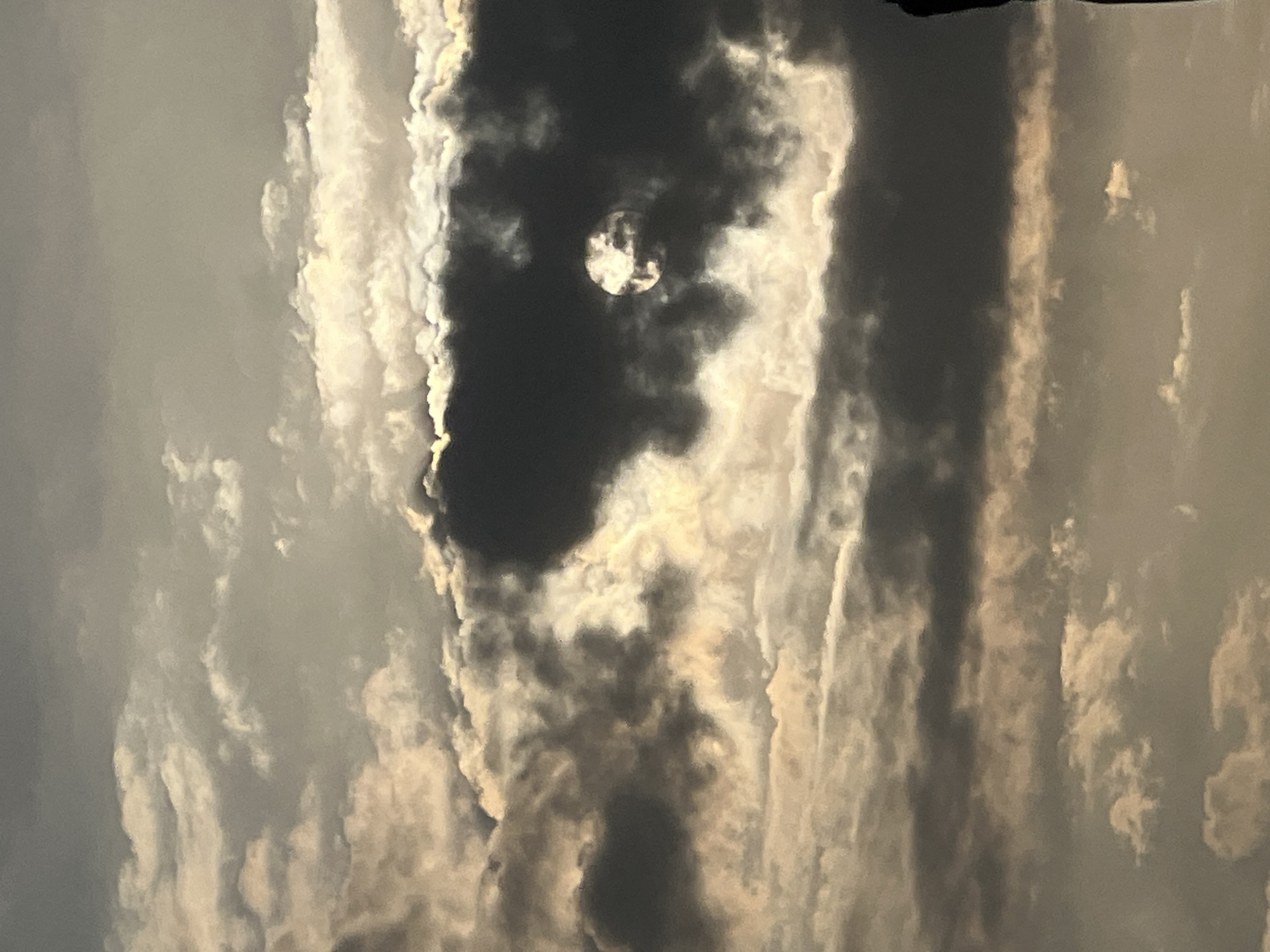 The image shows a view of the sky over Kuta, Indonesia, characterized by dramatic cloud formations. The sun is partially obscured by dark, heavy clouds, creating a sense of gloom and mystery. Lighter clouds surround the sun, catching its rays and creating a halo effect. In the lower right corner of the image, a dark silhouette of what appears to be a person or a structure is visible against the sky. The overall mood is somber, with the play of light and shadow on the clouds creating a sense of depth and texture. The scene is dominated by the sky, with no other discernible elements of the city visible.