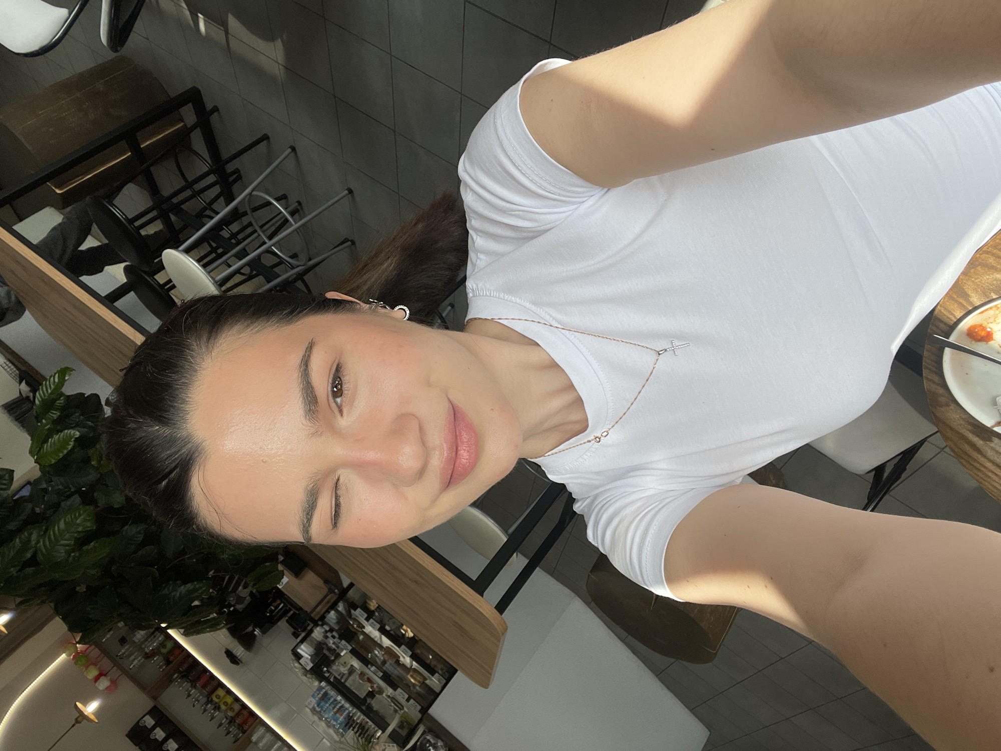 A woman takes a selfie indoors. She is wearing a white t-shirt and a silver necklace with a cross pendant. She is winking at the camera. Behind her, there are tables and chairs, as well as a shelf with various items on display. A plant is visible in the lower left corner of the frame. The location appears to be a café or restaurant, possibly in Krasnoyarsk, Russia, based on visual cues and the possibility of the image's origin.