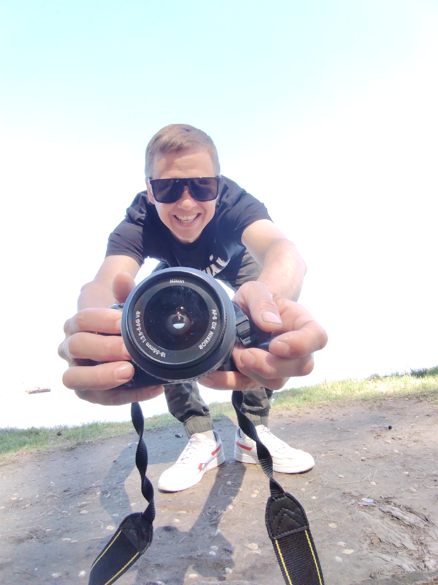 A man wearing sunglasses and a black t-shirt holds a Nikon camera towards the viewer. He is standing outdoors on a dirt path with white sneakers and jogger pants. The location is Chaikovsky, Russia. The sky is bright and almost white.