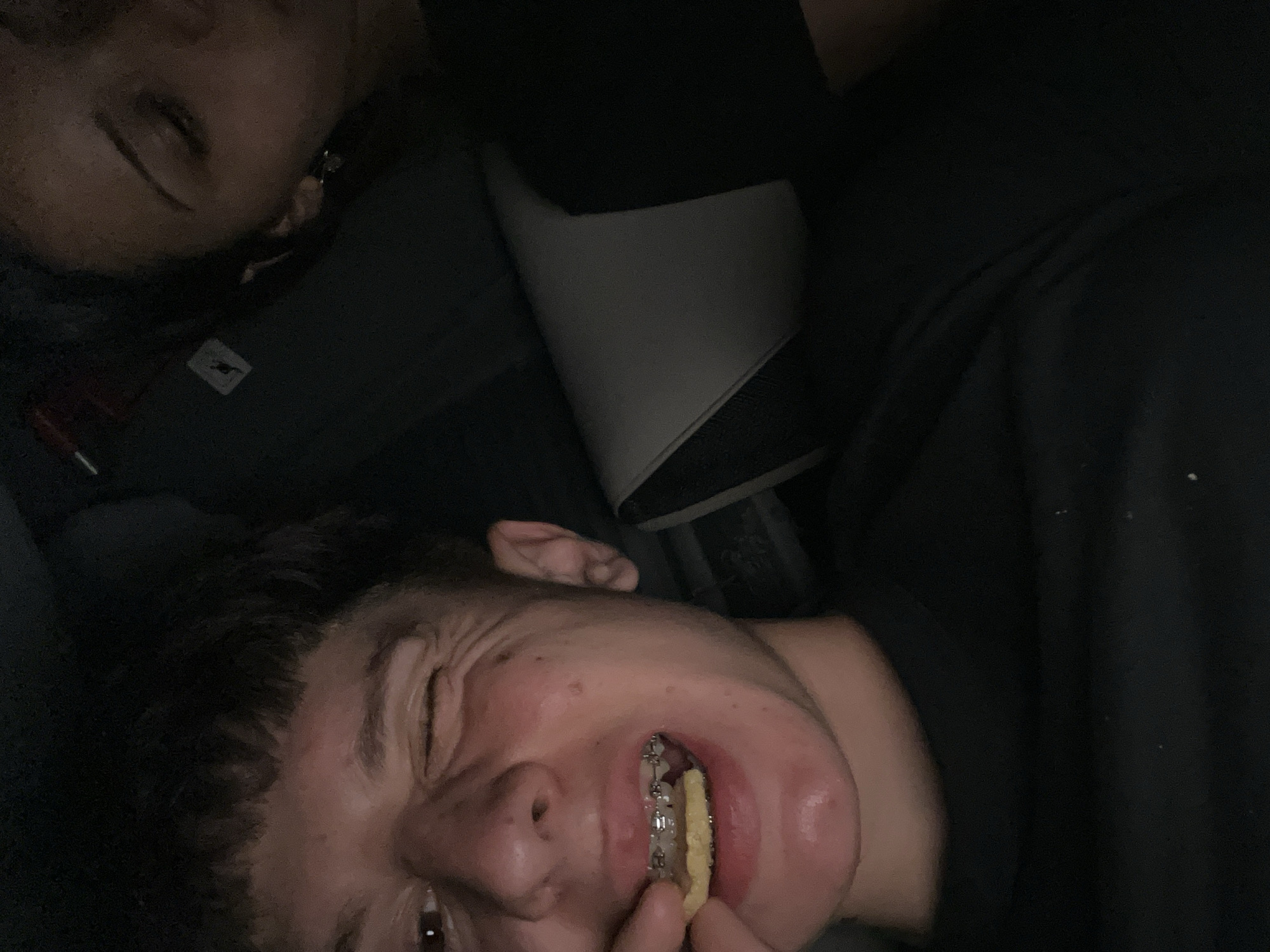 The image depicts two individuals lying down, possibly on a couch or bed. The person in the foreground is facing the camera, eyes squinted, and a piece of food is visible in their mouth, revealing braces. The person in the background is only partially visible, with their eye closed. The setting appears to be indoors, dimly lit. The location in the city of Popovitsa, Bulgaria is not discernible from the image.