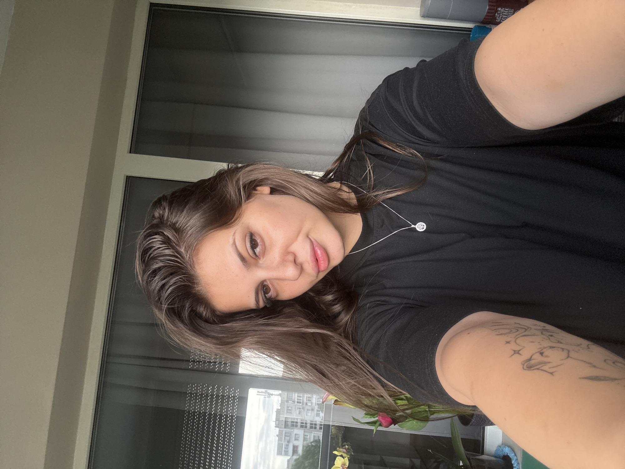A young woman with long, dark brown hair is taking a selfie in front of a window. She is wearing a black t-shirt and a silver necklace with a tree pendant. Her right arm is extended, and a tattoo of a cat can be seen on her forearm. Behind her, through the window, is a view of apartment buildings and trees, suggesting an urban setting. There are also some potted plants in the foreground, one with a pink flower. The lighting appears to be natural, coming from outside. The woman is looking directly at the camera with a slight smile. The scene is likely from an apartment or balcony in Plovdiv, Bulgaria, given the architectural style of the buildings visible.