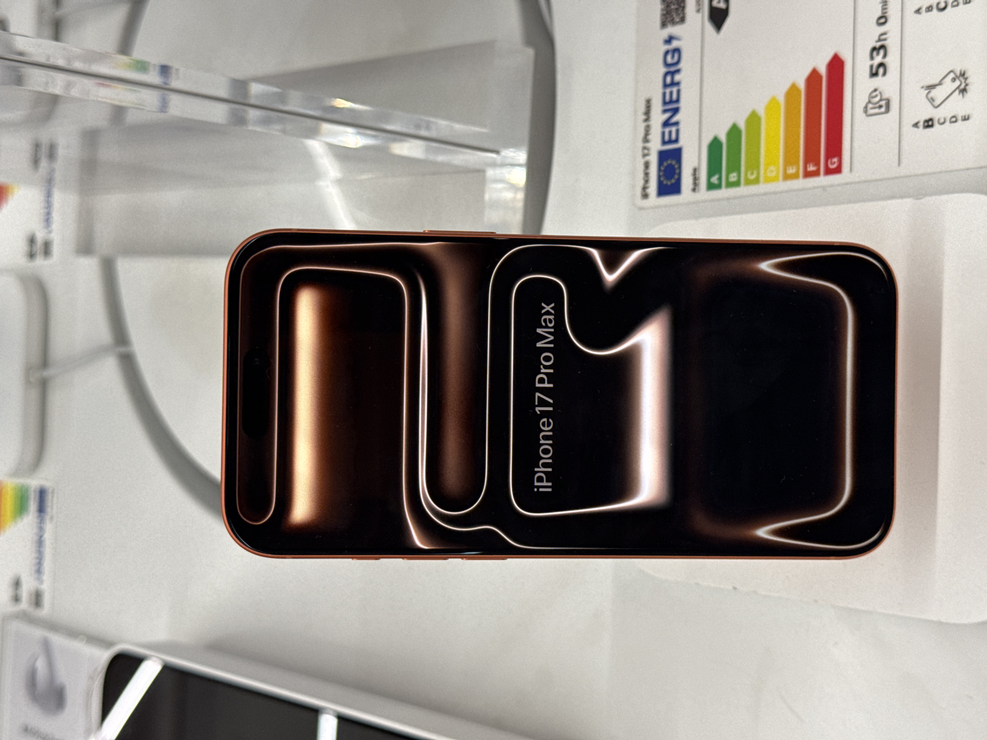 A smartphone, identified as an "iPhone 17 Pro Max," is prominently displayed in a retail store setting in Sofia, Bulgaria. The phone lies horizontally on a white surface, its screen activated to show a stylized dark background with flowing, glowing white lines, creating a sense of depth and movement. The model name "iPhone 17 Pro Max" is clearly visible in white text, rotated vertically on the screen. The visible frame of the device has a distinct reddish-brown or copper hue.
In the upper right portion of the image, an energy efficiency label is partially visible. The label prominently features the text "ENERG" and displays a standard European Union energy rating scale with colored bars ranging from A (green) to G (red). Further details on the label include "iPhone 17 Pro Max," "Apple," and "53h 0min," likely indicating battery performance or usage time. Transparent acrylic shelves or display elements are visible in the background, reinforcing the retail environment. The lighting suggests an indoor setting.