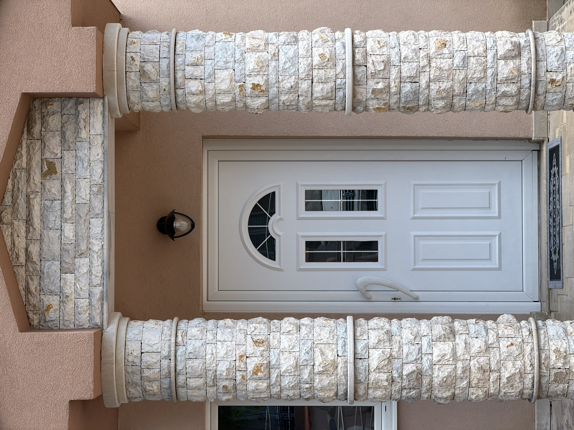 The image shows the entrance to a building with a light pink textured facade. Two cylindrical columns, constructed from stacked light-colored stones with visible pitting and occasional golden-brown inclusions, flank a white door. The columns are topped with a smooth, lighter stone cap and have a similar stone band encircling their base. Above the door, a section of the facade is covered with the same stone material as the columns, arranged in horizontal rows. A black outdoor lamp with a glass shade is mounted on the facade above the door. The white door has a semicircular arched top with a grid pattern and two rectangular windows below it, also with grid patterns. A white door handle curves upwards and to the left. To the left of the door is a white-framed window. A dark gray doormat with a lighter pattern is visible on the ground in front of the door. The scene appears to be an outdoor residential entrance in Radovići, Montenegro.