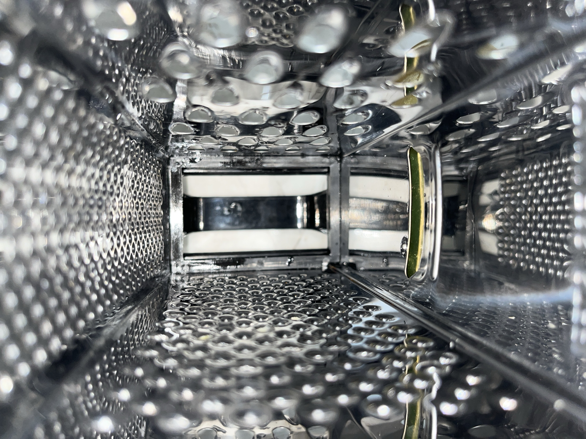 The image shows the inside of a metal box grater, angled upwards from the bottom interior. The grater surfaces are visible on all four sides, and some white and black rectangular objects are visible through the grating in the distance. The metal is shiny and reflective.