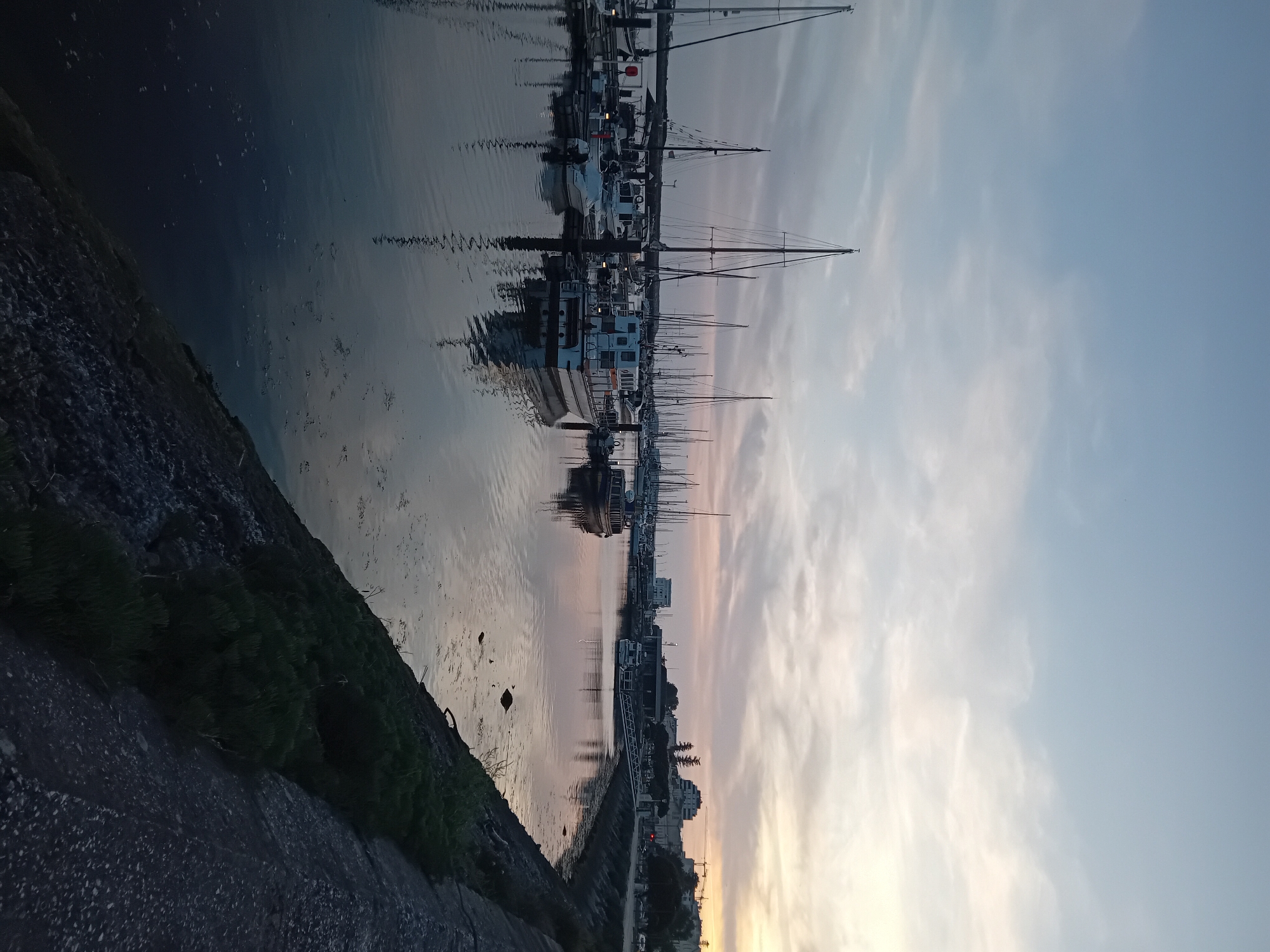 Captured at dusk, the image presents a view of a harbor in the city of Unknown, Unknown. The sky exhibits a gradient from light blue to hues of orange, signaling the setting sun. A harbor is populated with sailboats and motorboats, their masts vertical against the skyline. These boats are reflected in the still water, creating a mirror-like effect. Along the embankment, there's a paved walkway near buildings and sparse vegetation. In the foreground, a rocky wall descends to the water level. The illumination is dim, indicating that the scene is in the early stages of twilight.