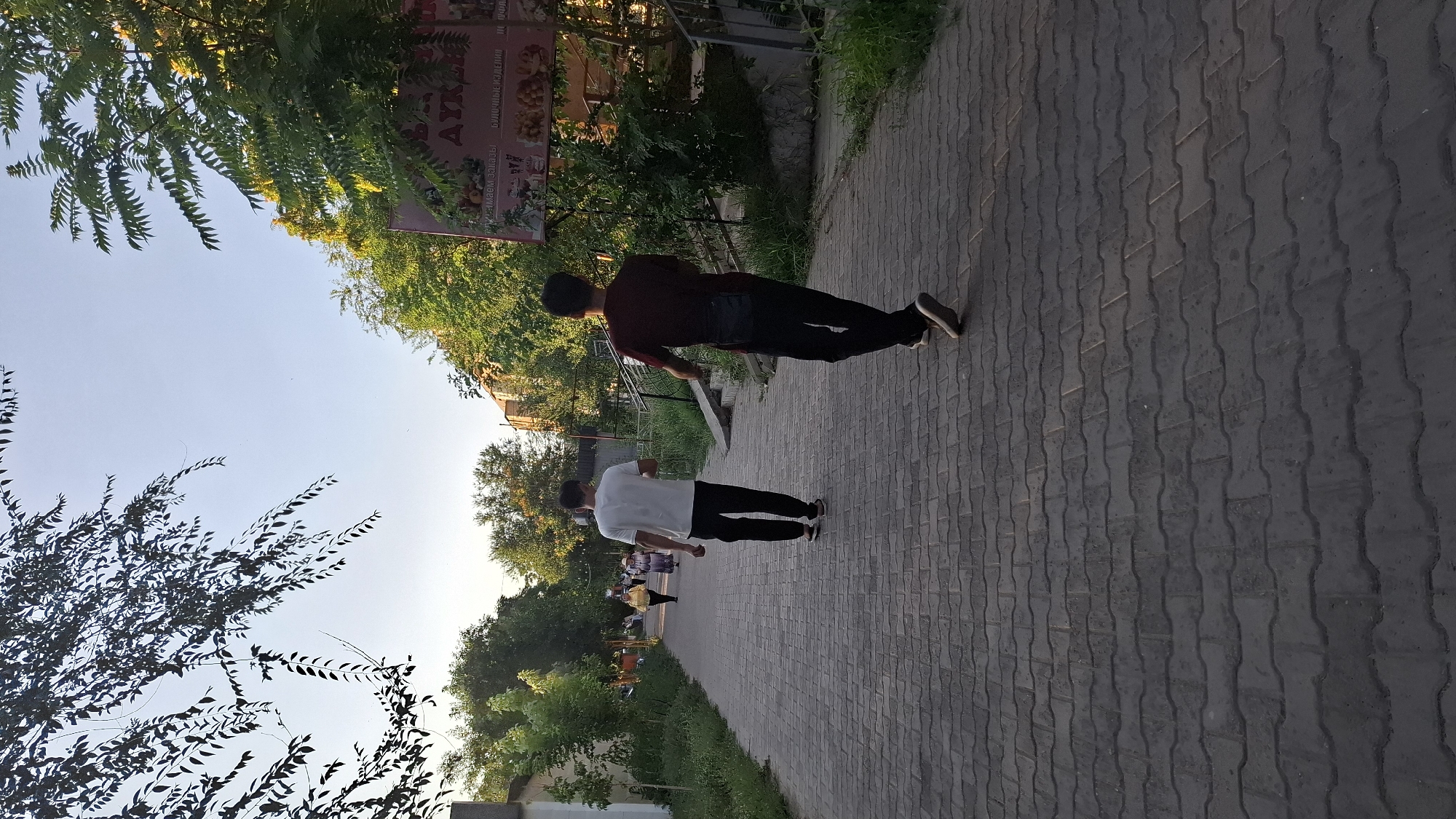The image shows a paved pedestrian walkway in an outdoor urban setting, likely in Shymkent, Kazakhstan, given the context. Two young men are walking away from the camera on the walkway. The man on the left wears a white t-shirt and black pants, while the man on the right wears a dark red t-shirt and black pants, with a small dark bag slung over his shoulder. In the background, several other people, including two women, are walking further down the path. Trees line both sides of the walkway, and buildings are visible behind the trees. A large, pink sign with text and images, possibly advertising a business, is also present on the right side of the frame. The overall lighting suggests it is late afternoon or early evening. The walkway is constructed with interlocking pavers.
