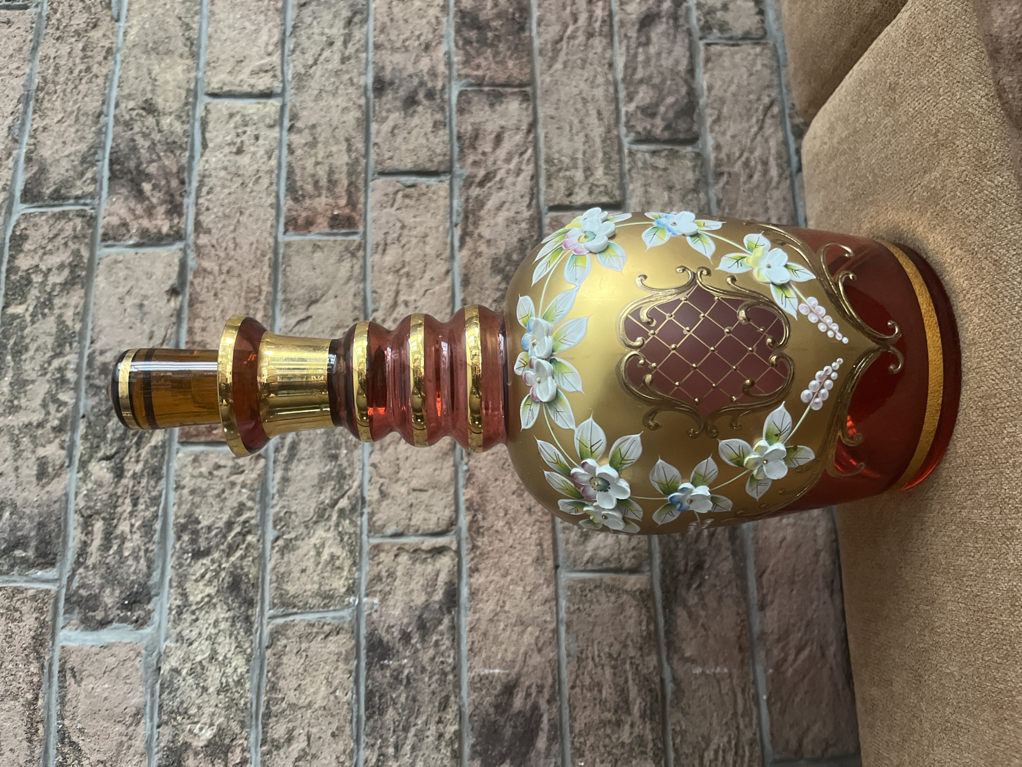 A decorative glass bottle with a gold-colored body and red accents is displayed against a brick wall. The bottle is adorned with white and blue floral patterns and features a decorative gold filigree design in the center. The bottle has a long, slender neck and a stopper. The background is a textured brick wall with visible mortar lines. The bottle is placed on a light brown, textured surface, possibly a piece of furniture. The lighting suggests an indoor setting, likely a home or display area. The image does not depict any people, actions, or interactions. No specific location in the city of Dnipro, Ukraine, can be identified from this image.