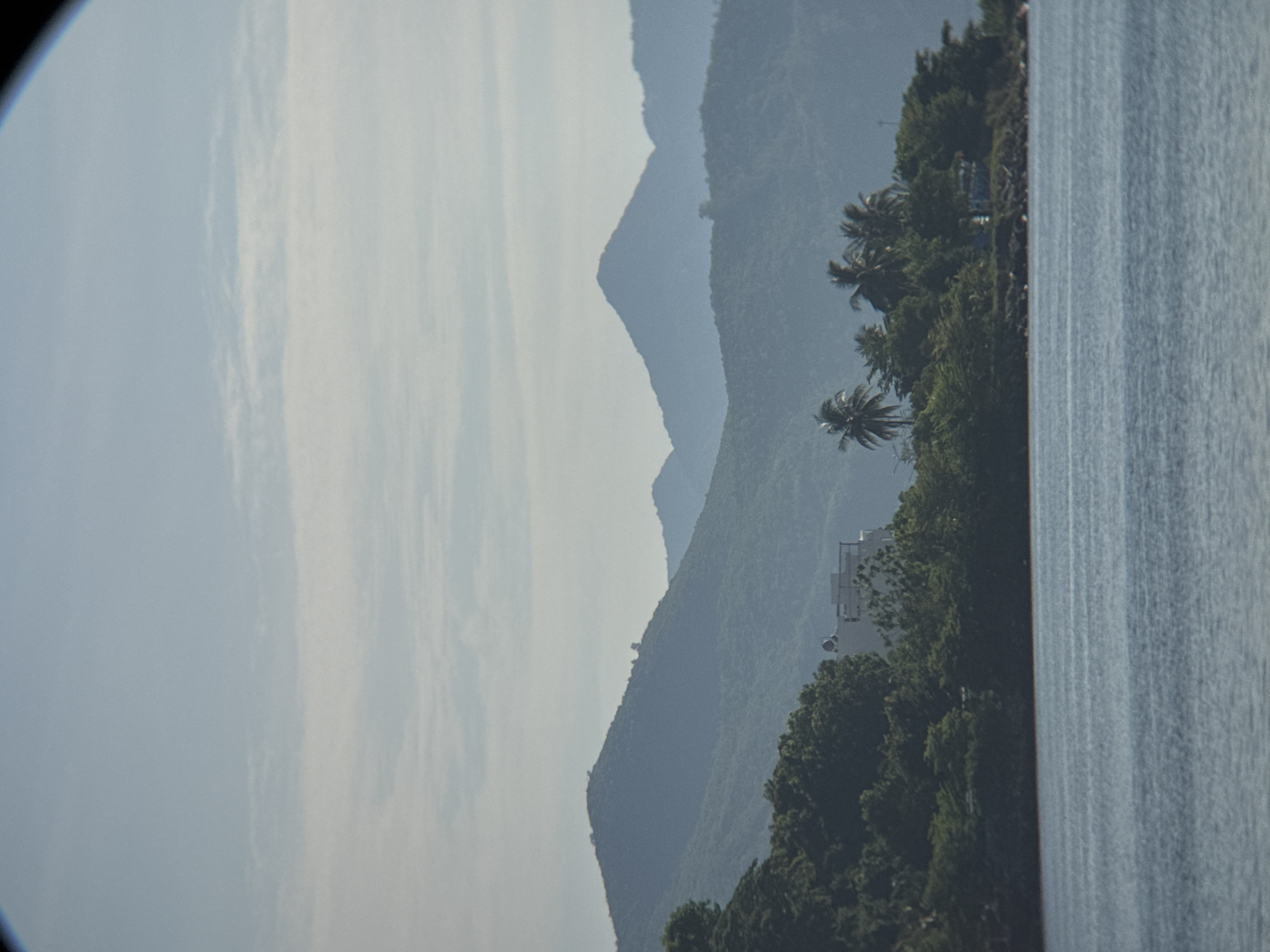The image depicts a tranquil coastal scene in Nha Trang, Vietnam, characterized by layered mountains receding into the horizon under a pale, cloudy sky. In the foreground, a body of calm, blue water occupies the bottom third of the frame. Along the shoreline, dense green foliage and scattered palm trees create a vibrant band of vegetation. A white building with a noticeable rooftop structure is partially visible amidst the trees on the left side of the image. The overall atmosphere is serene, with the natural landscape dominating the view.
