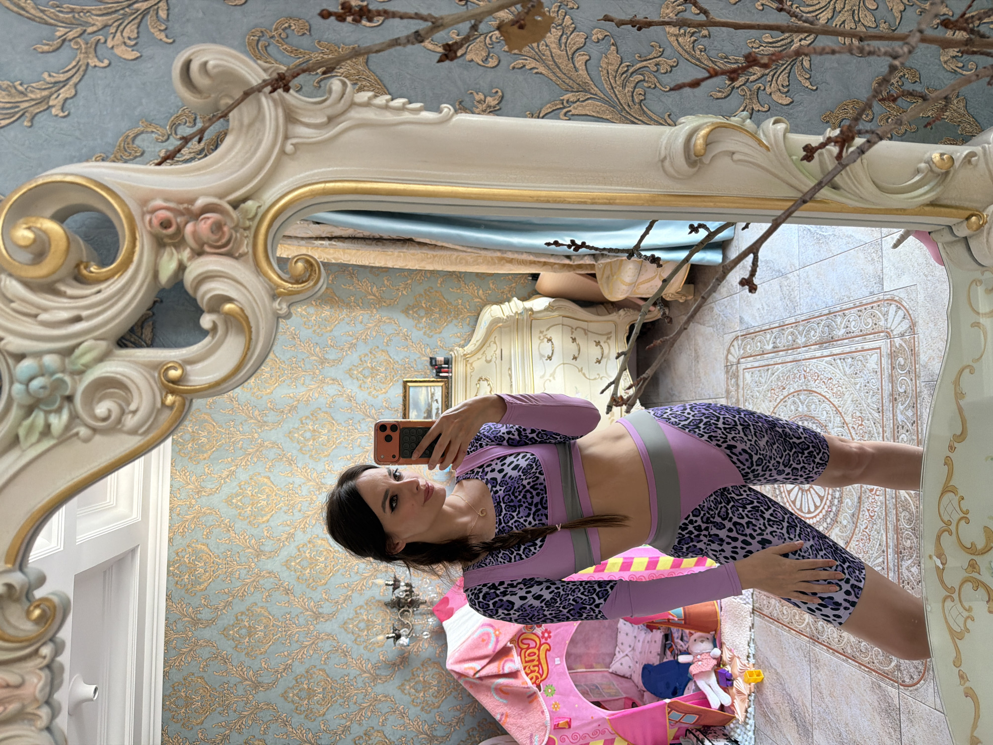 A woman is taking a selfie in a mirror in a room with ornate blue and gold wallpaper. She is wearing a purple and black leopard print athletic set with pink accents. She has dark hair in a braid and is wearing light makeup. In the mirror's reflection, a glimpse of a child's play tent, filled with toys, is visible. A large, ornate mirror with carved floral details sits on a dresser. The room appears to be a bedroom.