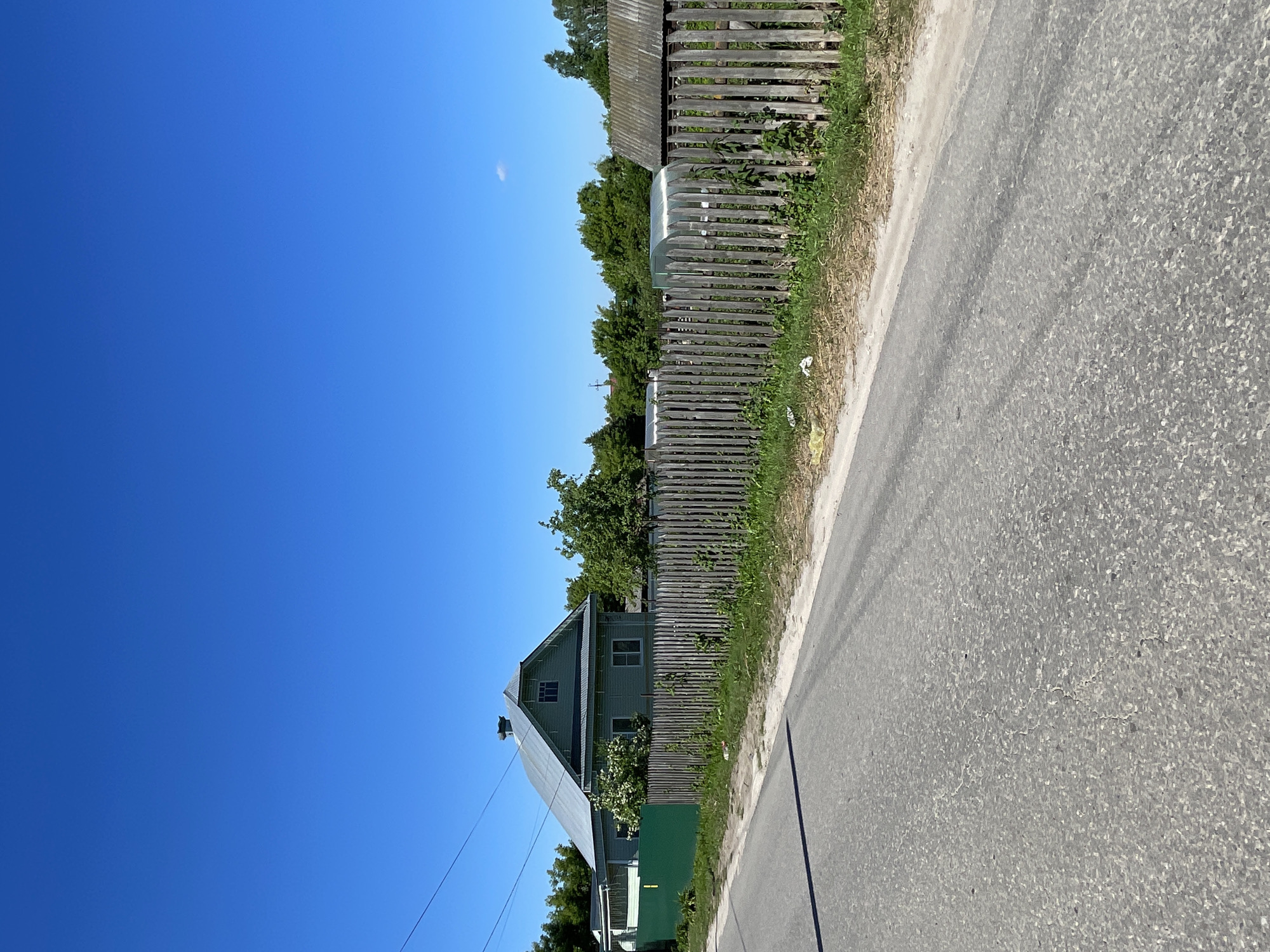 A rural scene in Krasnye Baki, Russia, under a bright blue sky. A paved road runs diagonally from the lower left to the upper right of the frame. Adjacent to the road is a wooden picket fence. Beyond the fence, a light green wooden house with a metal roof is visible. A dense line of trees forms the background, with some vegetation and a small greenhouse visible behind the fence to the right of the house. The scene appears to be a residential area in a village or small town.