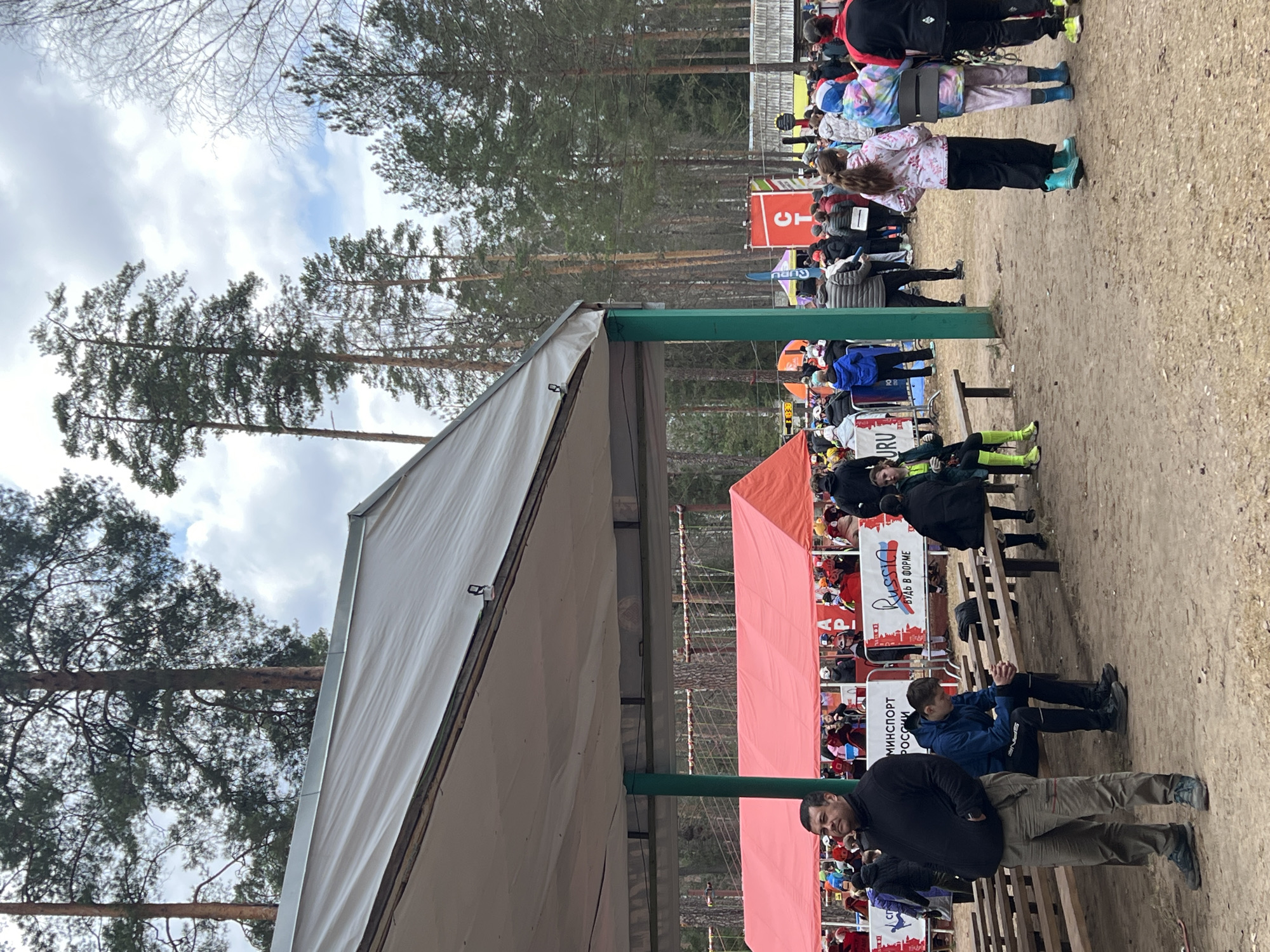 The image depicts an outdoor event taking place in a forested area of Lesnoye, Russia, under an overcast sky. The scene is populated by numerous people, several tents, and natural elements like pine trees and a dirt ground.
In the foreground and midground, groups of individuals are gathered. Many are dressed in warm, casual attire including jackets and long pants, indicating cool weather. Some people are standing, observing the scene, while others are seated on long wooden benches. A man in a black jacket and khaki pants stands prominently in the lower right. Another man in a blue jacket sits on a bench in the midground.
Two large tents are visible: a light-colored, possibly white or grey, tent dominates the left side of the frame, and a bright red tent is situated further to the right. A green vertical support beam stands between them. In the background, behind the red tent, an orange sign with partial Cyrillic text "СТ" is visible. Banners along the event area display text; one reads "RUSSIA" in English alongside the Cyrillic "ВУДЬ В ФОРМЕ" (BE IN SHAPE), and another below it states "МИНСПОРТ РОССИИ" (MINISTRY OF SPORT OF RUSSIA). These texts suggest the event is a sports or fitness-related gathering, likely endorsed by the Russian Ministry of Sport.
The background consists of a dense pine forest, with tall, green trees reaching upwards. Some bare deciduous branches are also visible against the cloudy sky, hinting at late winter or early spring. The ground beneath the attendees is unpaved dirt or sand. The overall impression is an organized outdoor community or sporting event.