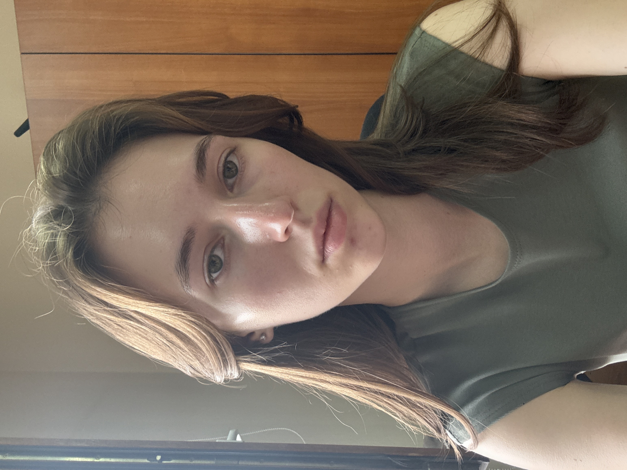 A young woman with long, brown, wavy hair is looking directly at the camera. She is wearing a dark green t-shirt. Her skin is fair and appears slightly shiny under the light. She has green eyes and a neutral expression. The background consists of a wooden cabinet or wardrobe on the right and a window frame with vertical blinds on the left. The scene is indoors, likely a room in a residential or office setting. There is no indication of the city of Perm, Russia in the image.