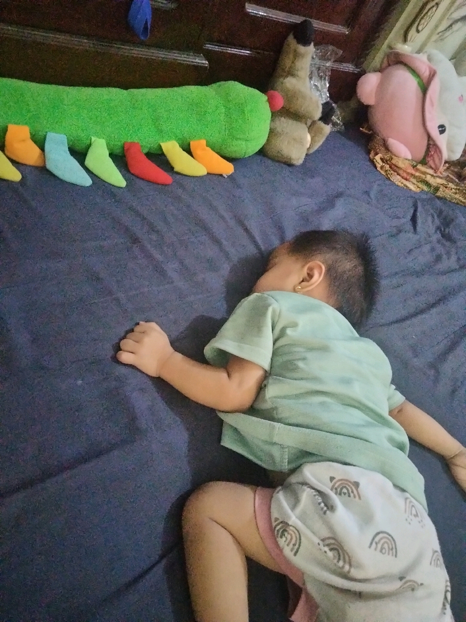 A young child is sleeping on a dark blue bed in Suka Sari, Indonesia. The child is lying on their side, wearing a light green t-shirt and light-colored shorts with a rainbow pattern. Their right arm is extended on the bed, and their left arm is bent. Several colorful plush toys are arranged on the bed near the child. These include a long, green caterpillar-shaped toy, a brown stuffed animal, and a pink stuffed animal with ears. The background shows a dark wooden surface, possibly furniture, and part of a patterned fabric.