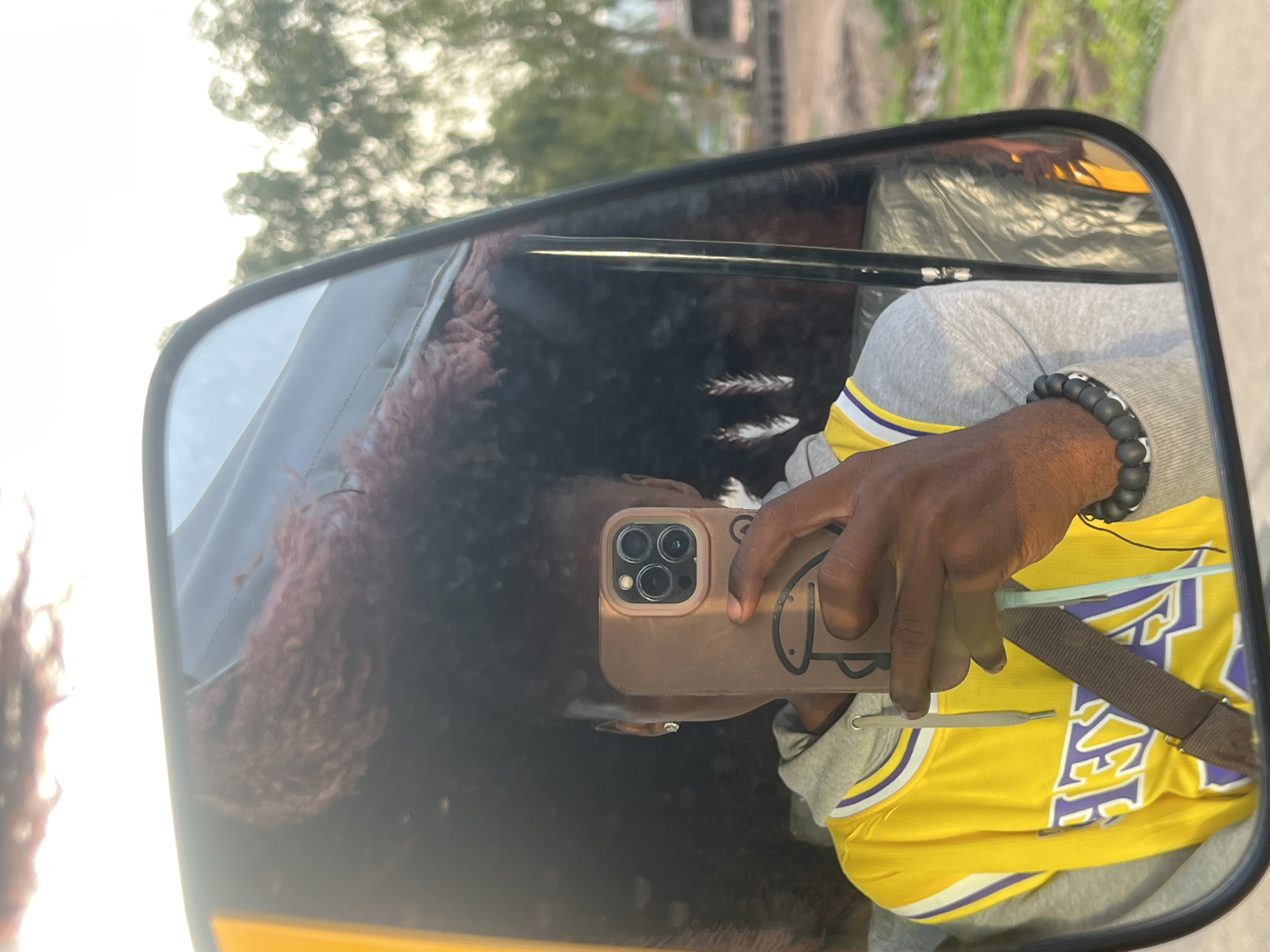 A person is reflected in a side mirror of a vehicle, likely a motorcycle taxi (keke), in Kporva, Nigeria. The person is holding a smartphone in a brown case with a cartoon face design, apparently taking a selfie in the mirror. The person is wearing a yellow Los Angeles Lakers jersey with purple trim, a gray hooded sweatshirt, and a black beaded bracelet. The reflection shows reddish-brown curly hair. The background includes blurred trees and buildings, suggestive of an urban environment, consistent with a location in Kporva. A seat belt crosses the person's chest.
