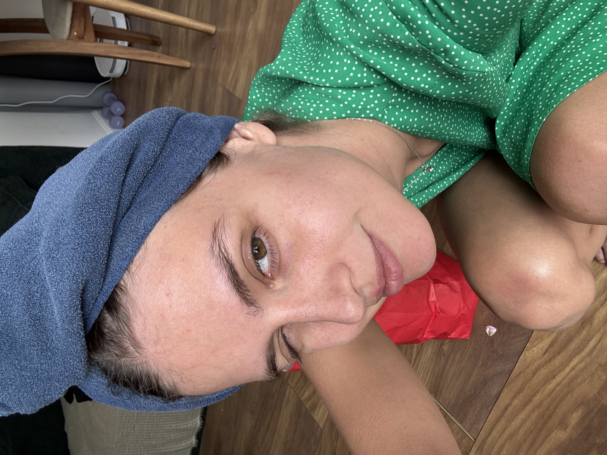 A person is lying on a wooden floor, wearing a green polka dot dress and a blue towel wrapped around their head. A red object is near their face. Parts of furniture are visible in the background. The location is Unknown, Unknown.