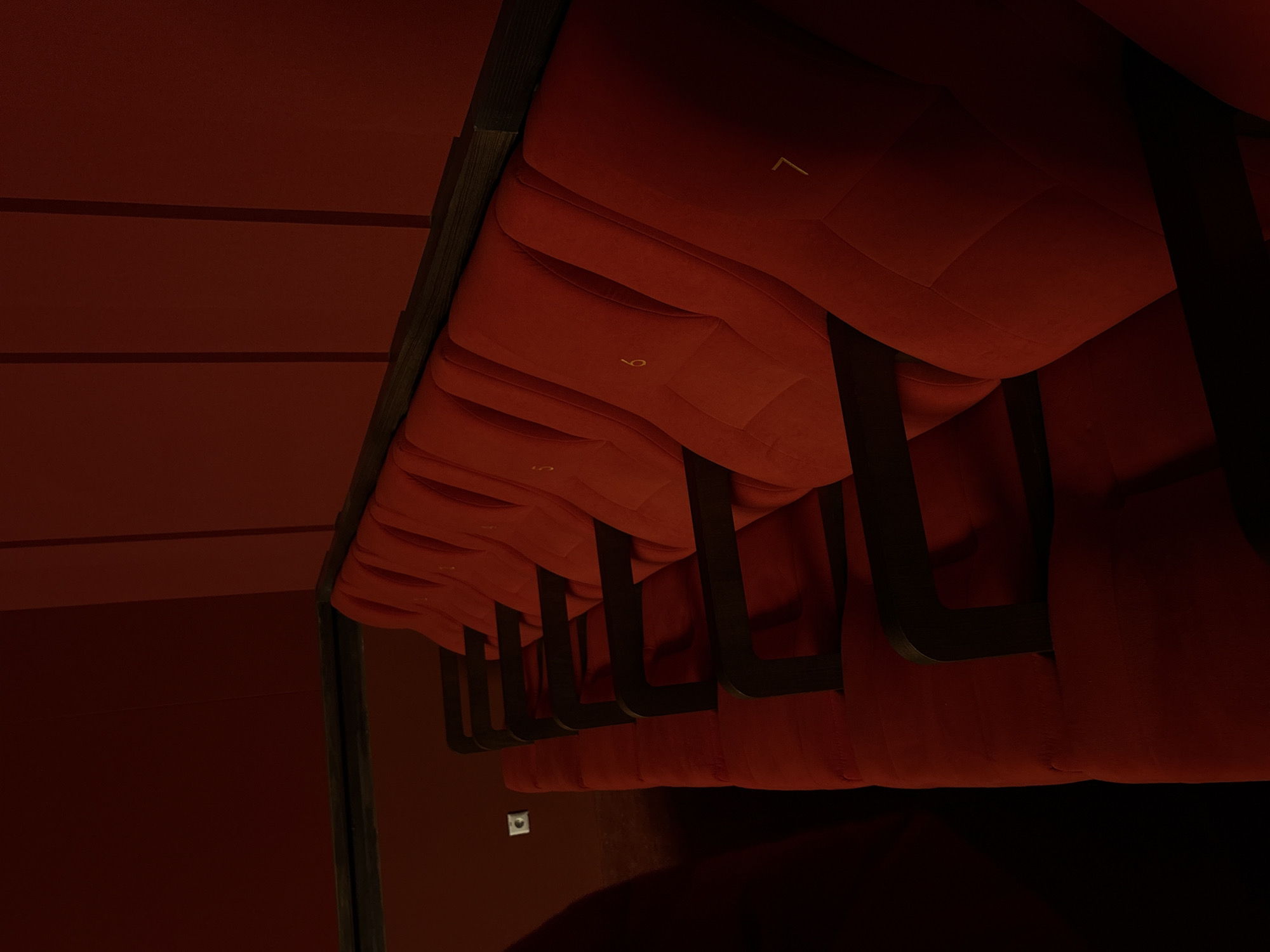 The image depicts a row of red cushioned theater seats in a dimly lit room. Each seat has a number on the cushion, ranging from "7" to "5". The backrests have a textured, pleated design. Dark-colored armrests separate each seat. The ceiling and walls are a similar shade of red. The location cannot be determined from the image. The image does not provide any information regarding subjects, objects, actions, or interactions. The image is an interior shot of a seating area, likely in a theater or auditorium.