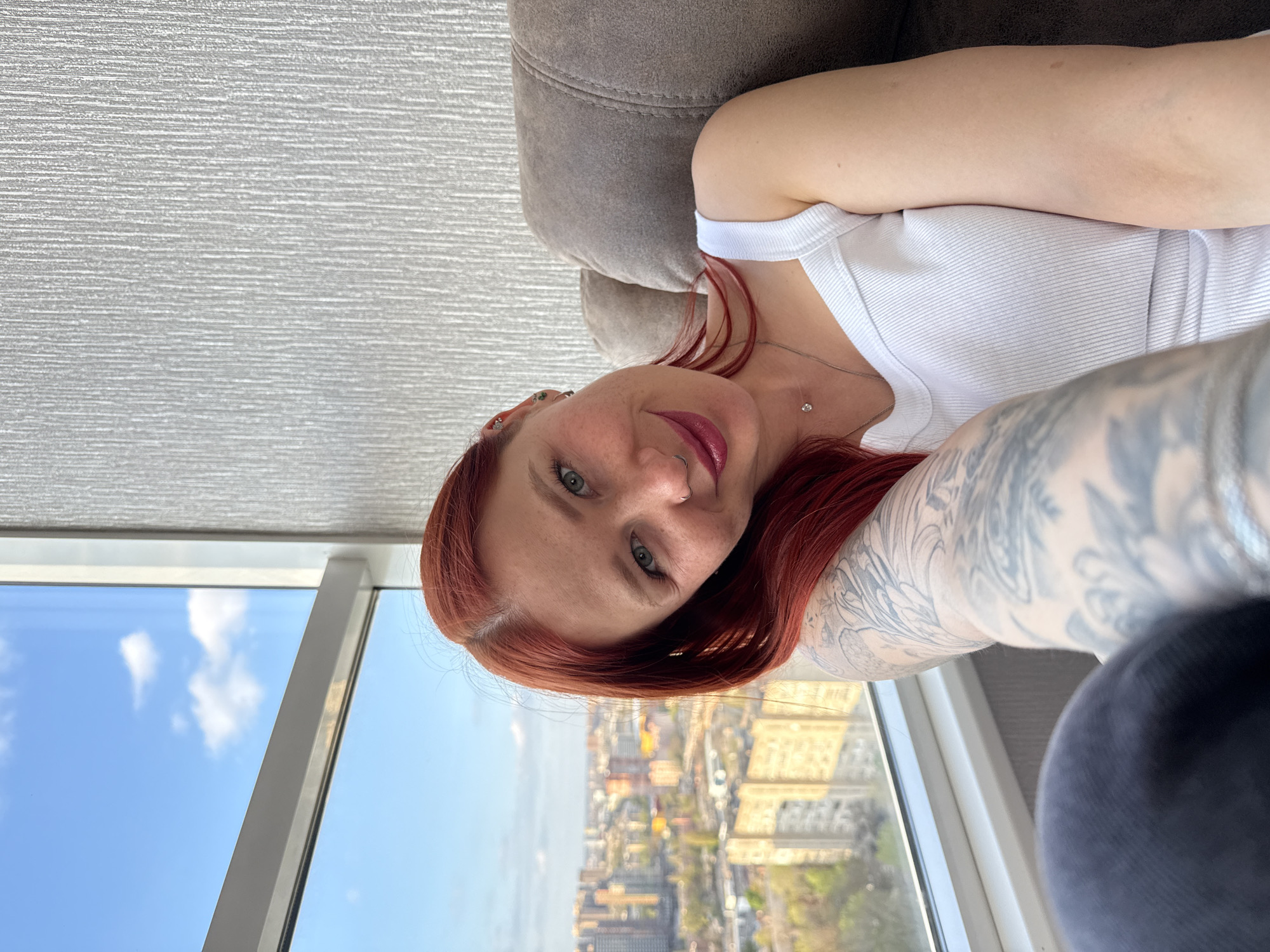 A selfie portrait captures a woman indoors, looking directly at the camera with a subtle smile. She has long, reddish-auburn hair, green eyes, and fair skin. Her lips are adorned with dark pink or berry-colored lipstick. Visible facial piercings include a silver nose ring and multiple silver hoops and studs in her left ear. She wears a simple white ribbed tank top and a delicate silver necklace with a small, clear gemstone pendant. Her right arm, prominent in the foreground, is covered with extensive grey and black tattoos, forming a detailed sleeve.
The woman appears to be reclining against a textured gray sofa or cushion, which is visible in the upper right quadrant of the image. Behind her, a large window offers a panoramic view of a city under a bright blue sky with scattered white clouds, indicating daytime. The cityscape features a mix of buildings, some with ornate architecture and others more modern, extending into the distance. The background wall on the left side of the frame has a textured light gray or white finish.
The overall scene conveys a casual and relaxed moment, capturing the woman against an urban backdrop, consistent with a setting in a large city like Rostov-on-Don, Russia. No visible text is present in the image.