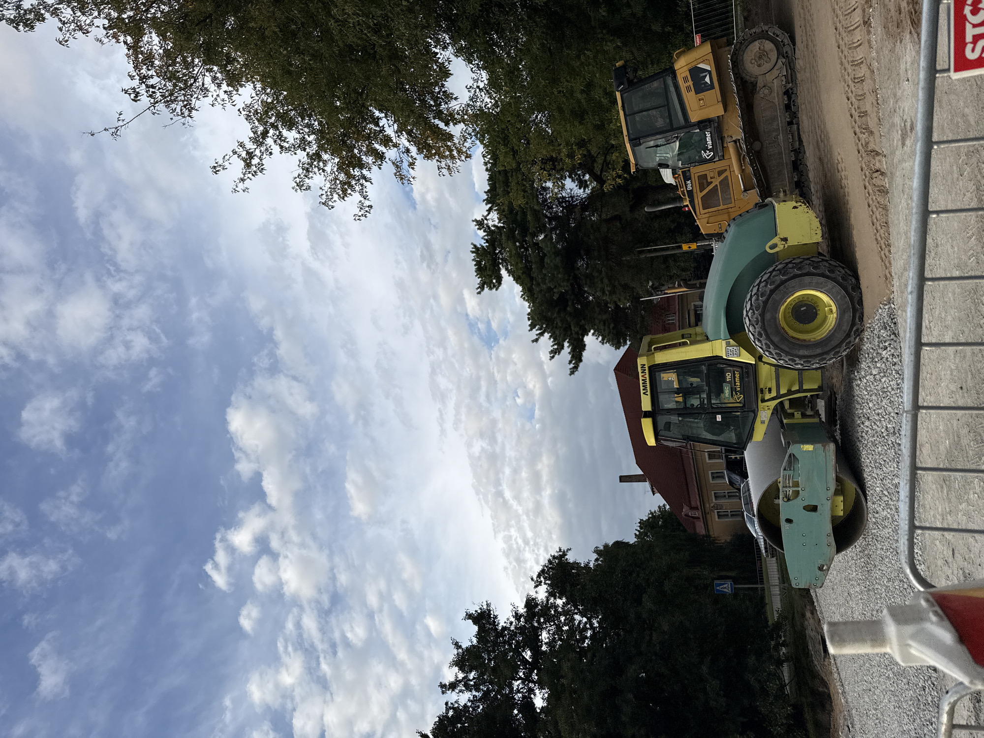 The image shows a construction site in Tallinn, Estonia, with two pieces of heavy machinery: a yellow and green vibratory roller on the left and a yellow bulldozer on the right. The roller is positioned on a surface covered with gravel, while the bulldozer sits on a sandy, excavated area with tire tracks. In the background, a building with a red roof is visible, surrounded by trees. The sky above is partly cloudy, with a mix of blue sky and white clouds. A pedestrian crossing sign is present to the left of the roller. A metal barrier runs across the foreground of the image.