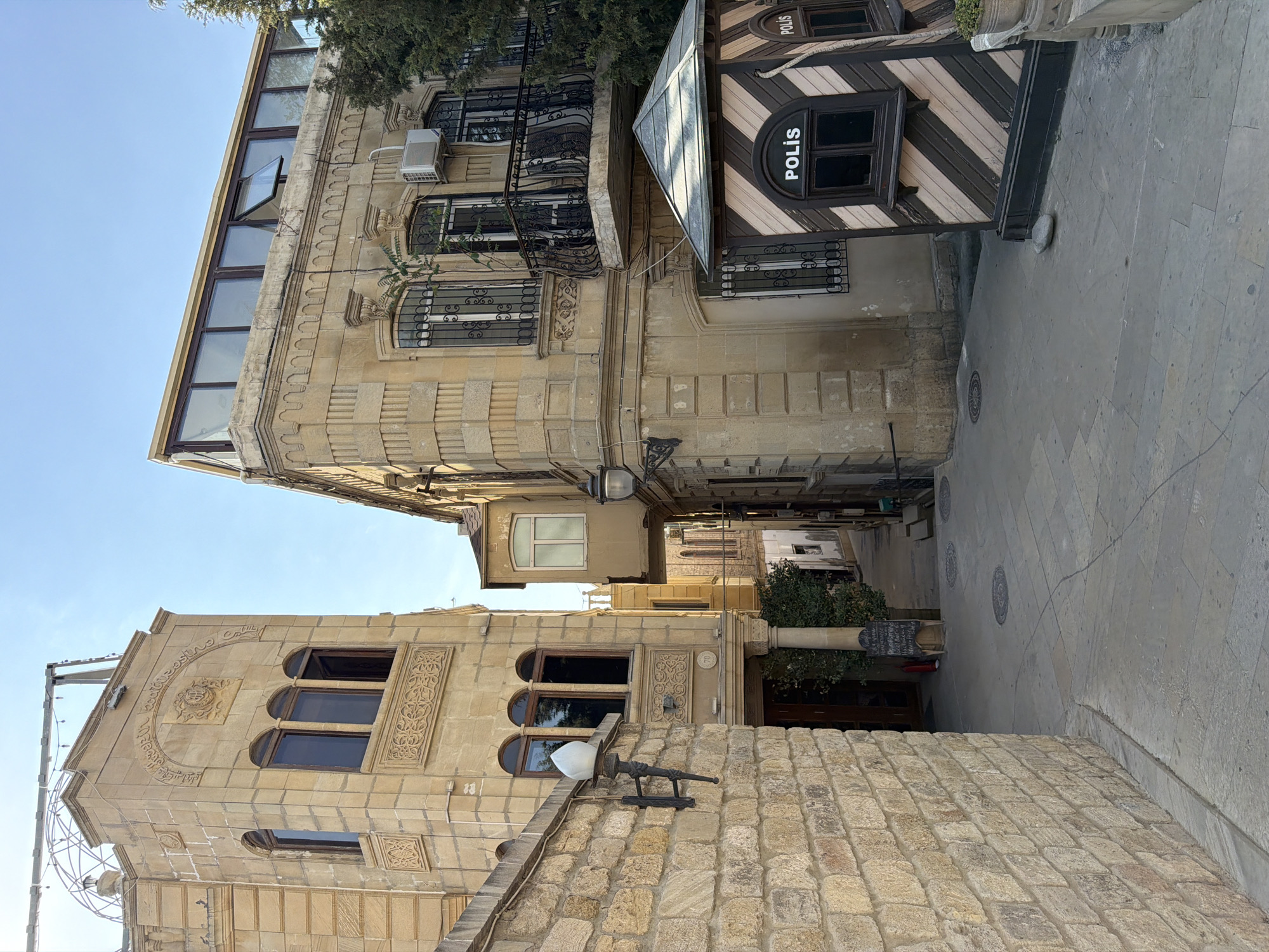 A narrow, cobblestone alleyway is depicted in the historic Old City of Baku, Sabail Raion, Azerbaijan. Two buildings, constructed with light-colored stone, flank the pathway. The building on the left is a multi-story structure with arched windows and ornate carvings. The building on the right has a more modern addition on its upper level, featuring a large, glass-enclosed space. In the foreground, a small, dark wooden booth with the word "POLİS" visible on its windows stands to the right. The sky is clear and blue. The scene appears to be under natural daylight.