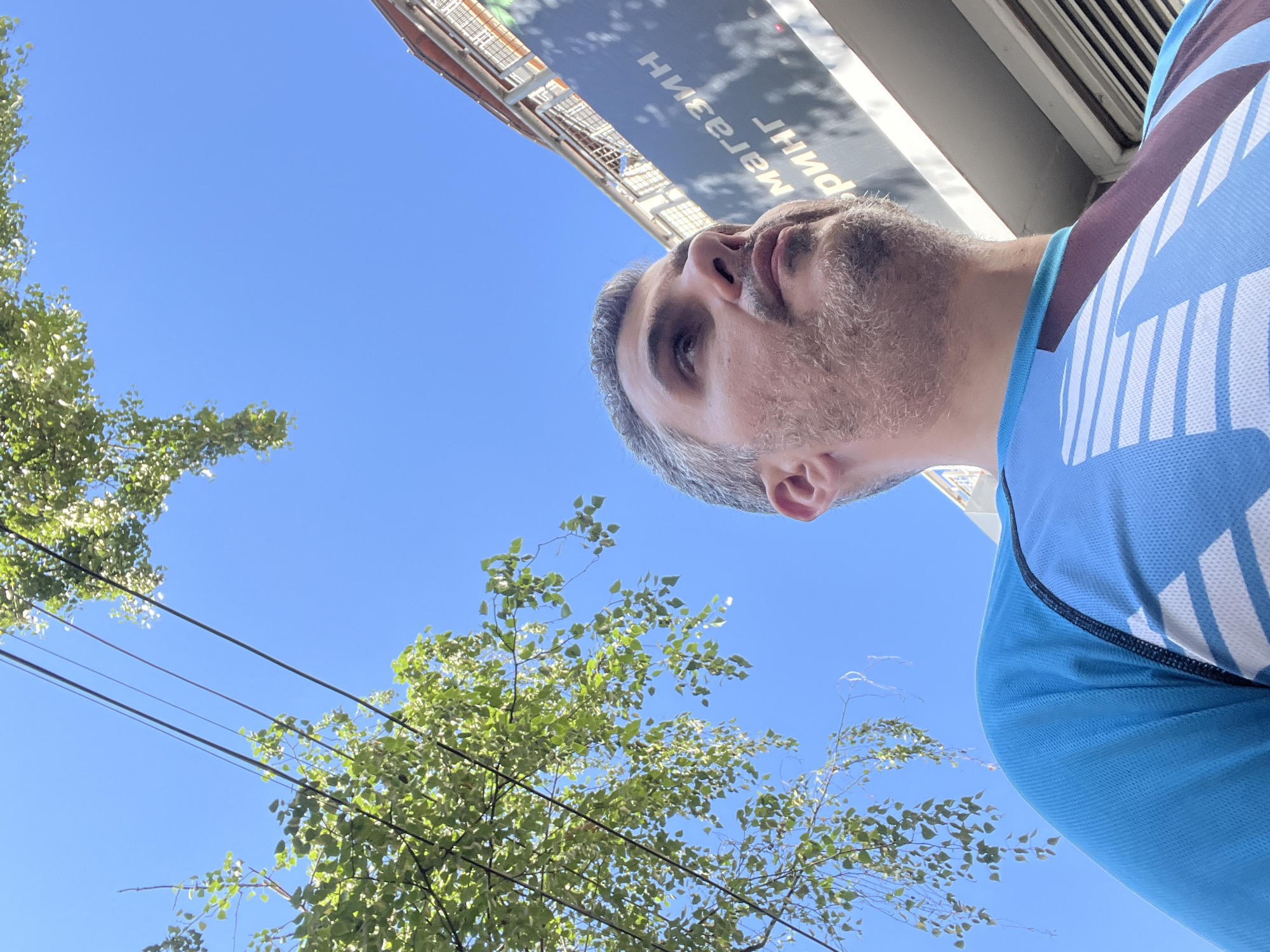 A medium shot captures a man in a blue and white patterned athletic shirt, viewed from a low angle looking up at the sky. The man has graying hair and a beard, and he is looking upwards and to the right. To his left, a tree with green leaves hangs into the frame, with power lines visible against the clear blue sky. Behind him, a building with a sign is partially visible. The scene appears to be outdoors in a city.