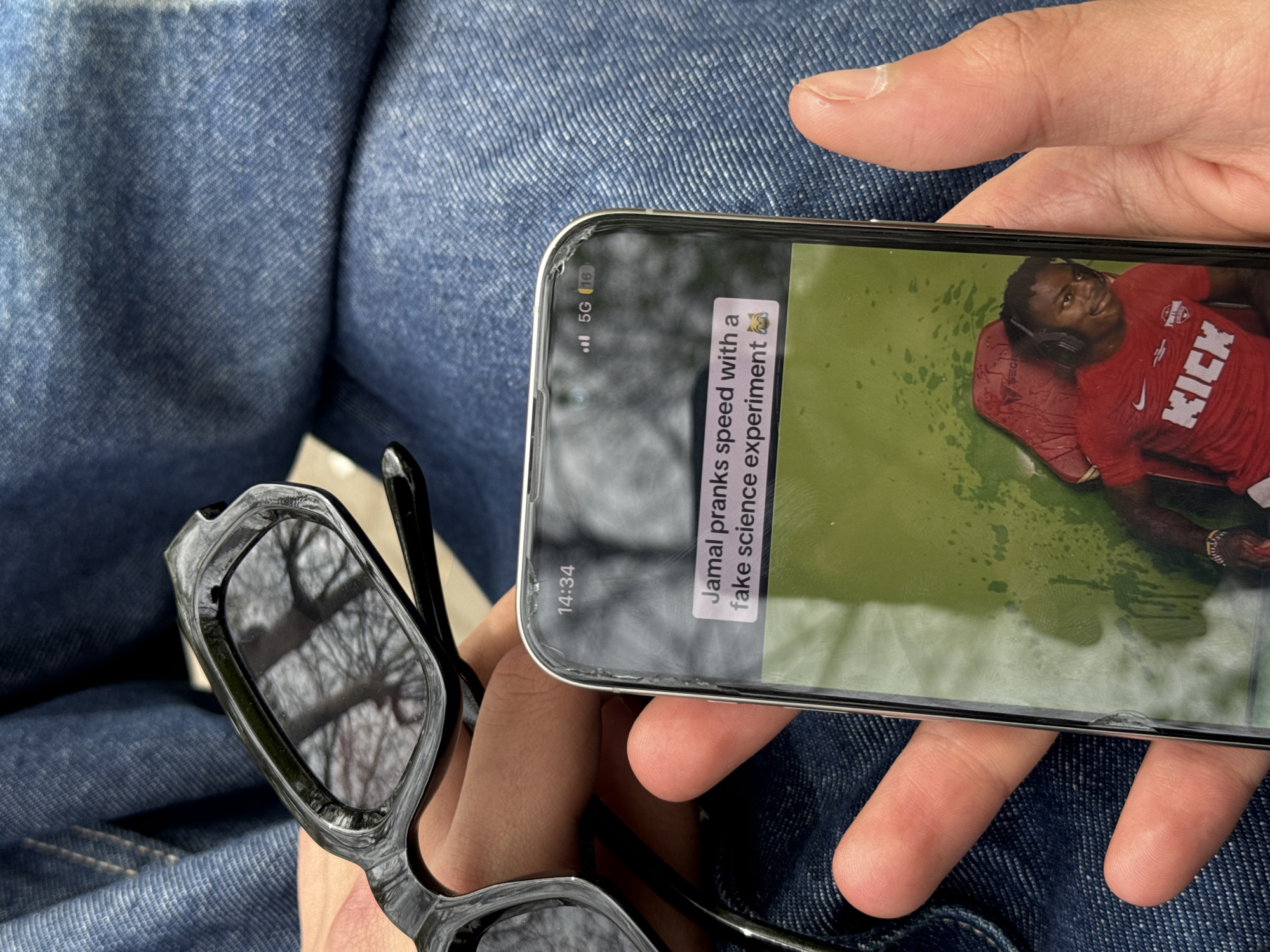 A person is holding a black iPhone with a cracked screen displaying a video of a person in a red shirt that reads “KICK” with the text “Jamal pranks speed with a fake science experiment.” The iPhone is in the person’s right hand, and they are also holding black-rimmed sunglasses in their left hand. The person is wearing blue jeans. The reflections in the sunglasses show bare trees, suggesting an outdoor setting. The city and location of Yerevan, Armenia, are not identifiable from the image.