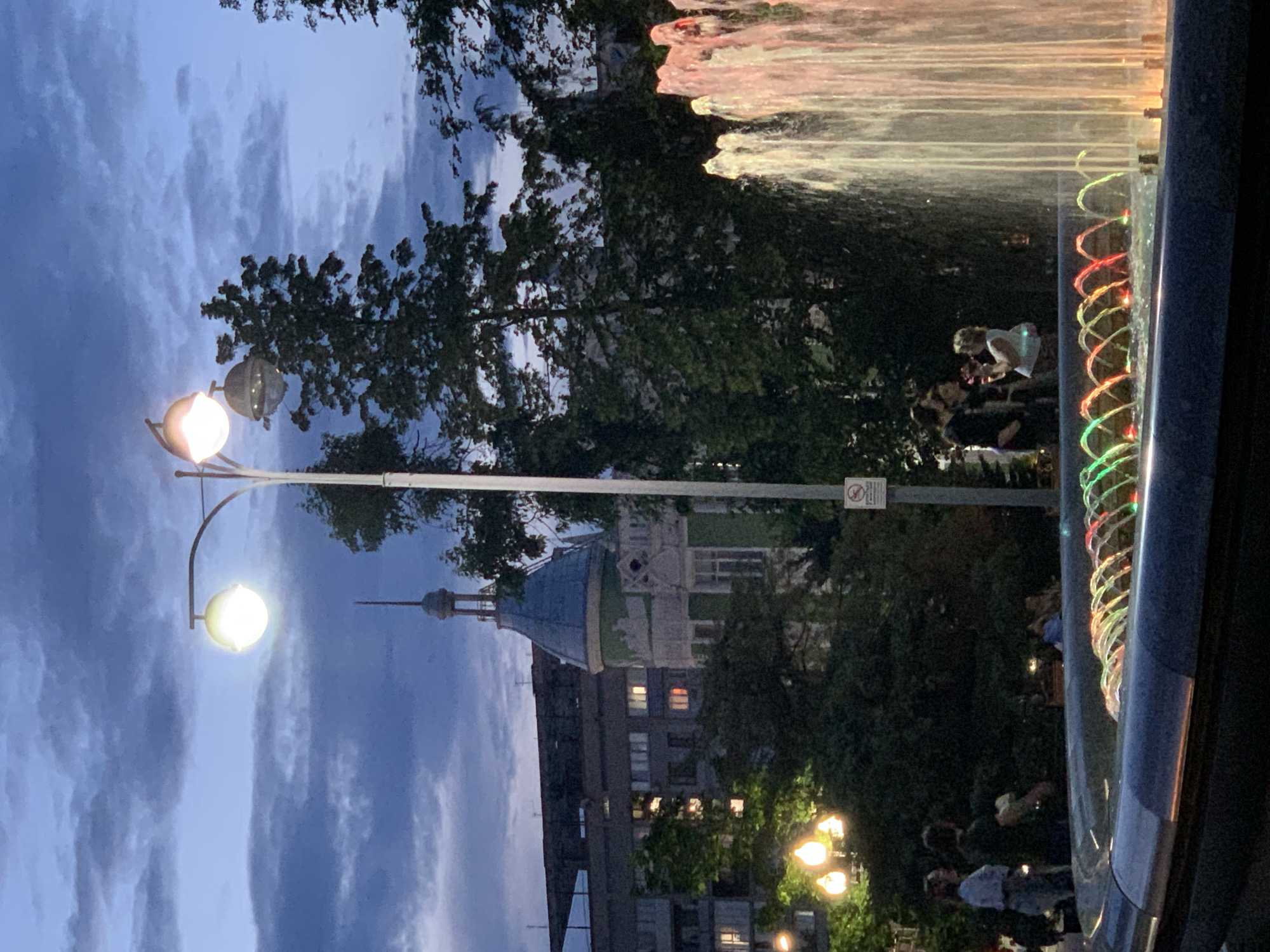In the city of Unknown, Unknown, this image captures an evening scene by a fountain. The sky is a deep blue with clouds, suggesting twilight or a cloudy evening. A streetlamp with two globes illuminates the area. In the background, a building with a distinctive green turret and spire is visible, along with other urban structures. Trees are interspersed throughout the scene, some with lush green foliage. A fountain with colorful, illuminated water jets is in the foreground. Several people are present, some standing near the fountain, and others further back, appearing to interact or observe the fountain. One person is holding a small object, possibly a phone, and another appears to be a child. The overall atmosphere is that of a public urban space at dusk.