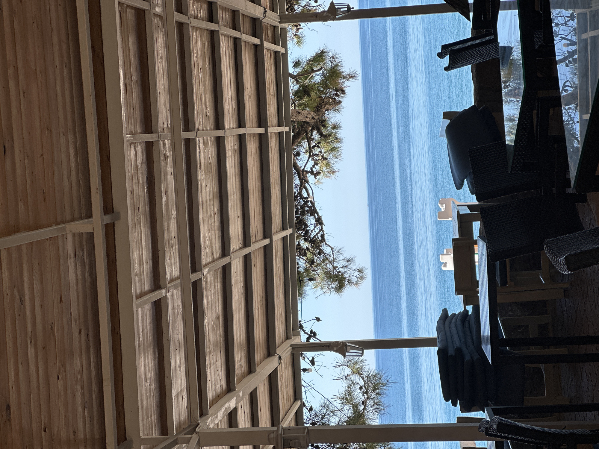 View from an outdoor seating area, possibly a restaurant or cafe, in Boreti, Montenegro. The camera looks out over the Adriatic Sea under a wooden-slatted awning. The interior of the seating area includes tables and chairs, stacked cushions, and structural supports. Through the open space beyond the awning, the sea stretches to the horizon under a clear sky. A few boats are visible on the water. Trees are visible on the side.