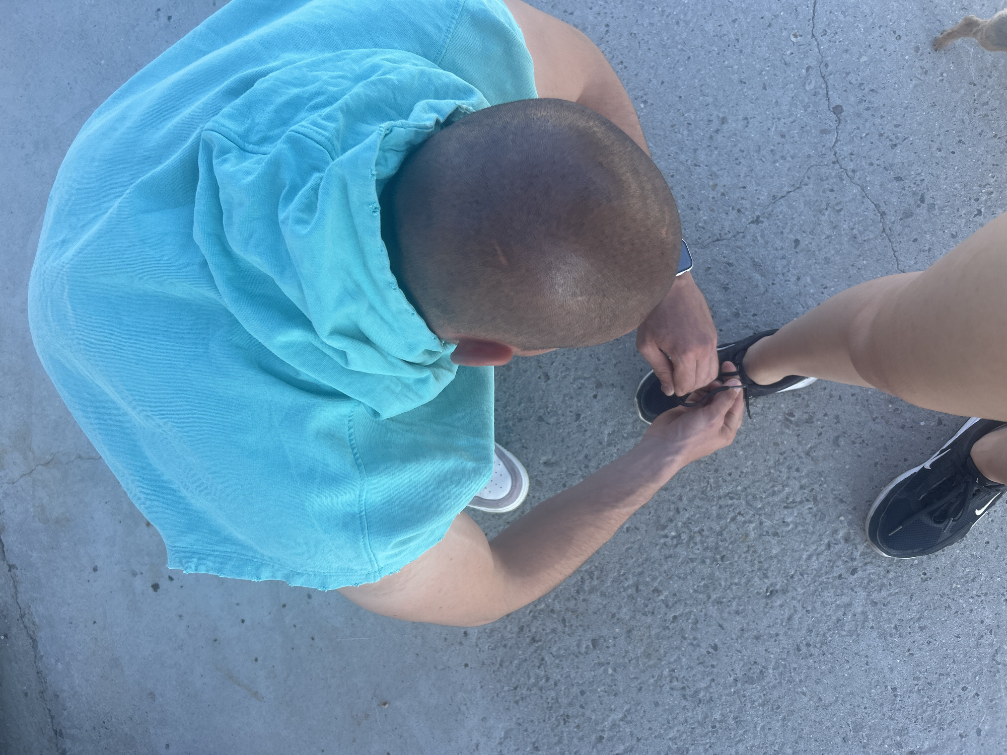 A high-angle shot depicts a man tying the shoelace of a person standing next to him on a grey concrete surface, likely outdoors. The man has a shaved head and wears a light blue hooded t-shirt. He is bent over, his arms extending towards the other person's foot. The person he is assisting is wearing black Nike sneakers. The background consists of a textured grey concrete surface with varying tones. The location is uncertain, though not defined in this context.