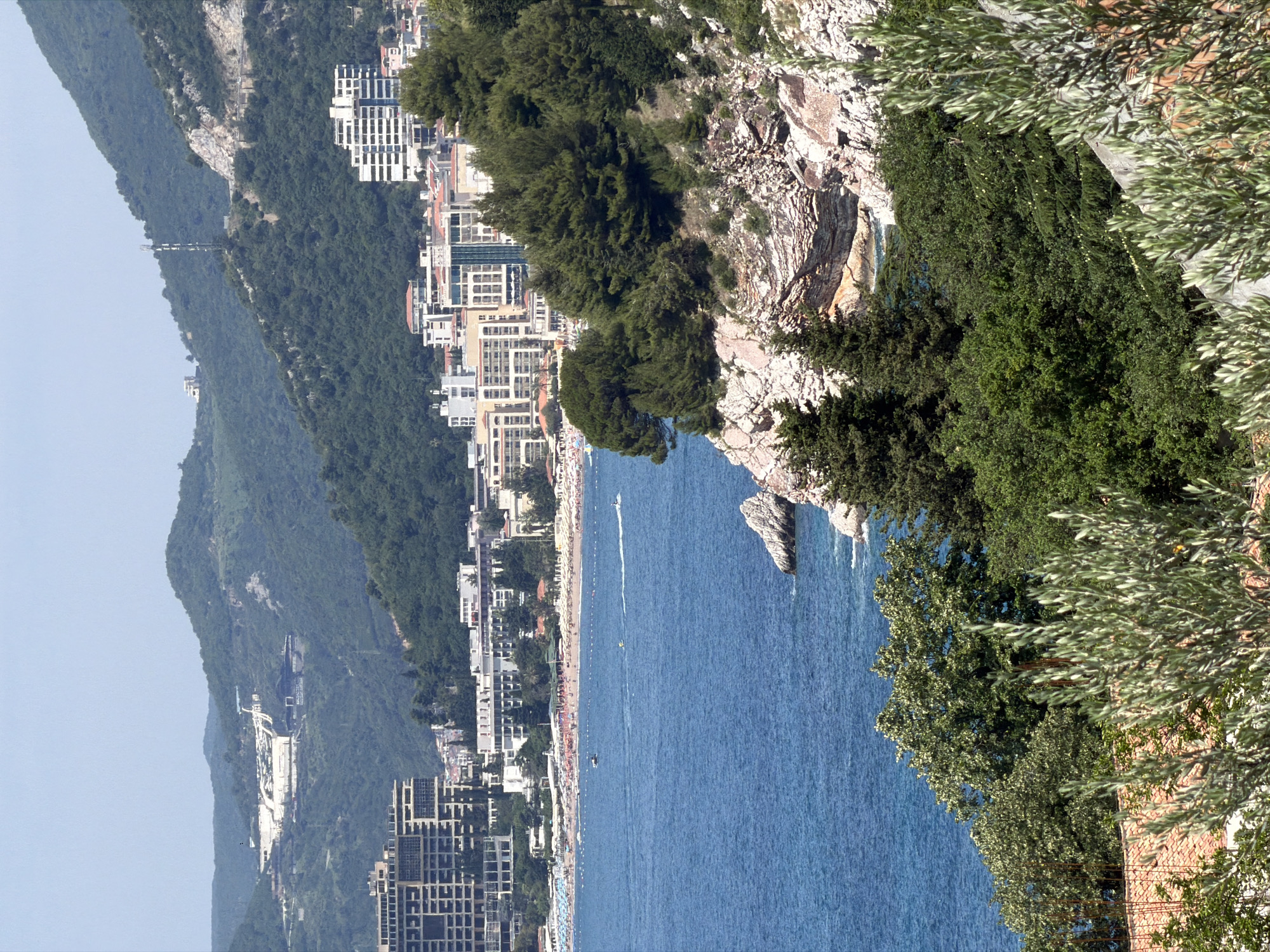 The image depicts a coastal view of the city of Vrijesno, Montenegro. In the foreground, a rocky cliff face is covered with dense green vegetation, including olive trees and evergreen trees. Below the cliffs, the azure waters of the Adriatic Sea are visible, with a rocky outcrop protruding from the water on the right side. In the mid-ground, a sandy beach is lined with numerous beach umbrellas and a few scattered beachgoers. Beyond the beach, a dense collection of buildings, consisting of hotels and apartment complexes with red-tiled roofs and white facades, stretches along the coastline. Further inland, steep, tree-covered hills rise, with more modern buildings visible higher up on the slopes. A telecommunications tower is perched on one of the hilltops. The sky is clear and a pale blue.
